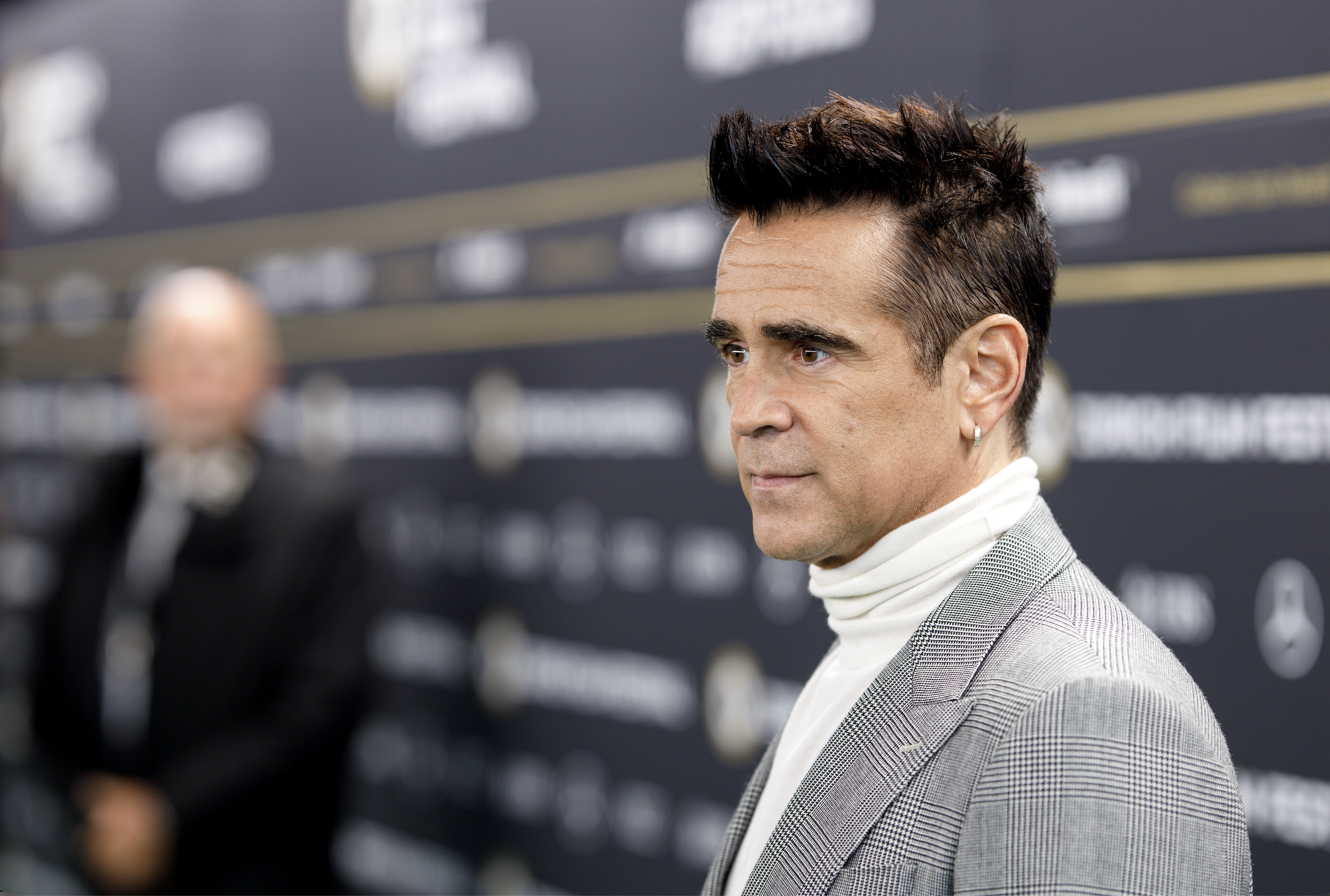 A man with dark hair styled back, wearing a light gray checkered blazer and a white turtleneck, at a formal event with a black backdrop and company logos.