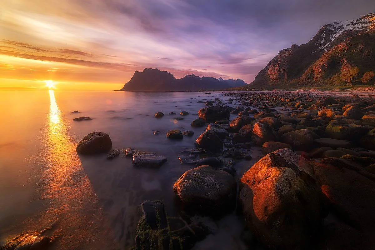 Sunset over rocky shoreline with mountains in the background and colorful sky.