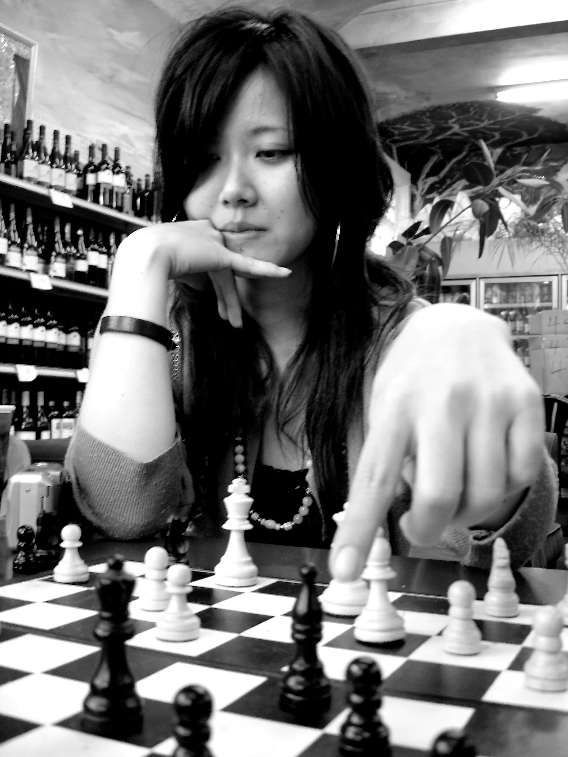A woman with long dark hair plays chess in a store or cafe, focusing on her next move. She wears a bracelet, a necklace, and hoop earrings. Shelves with wine bottles and calendar boxes are visible in the background.