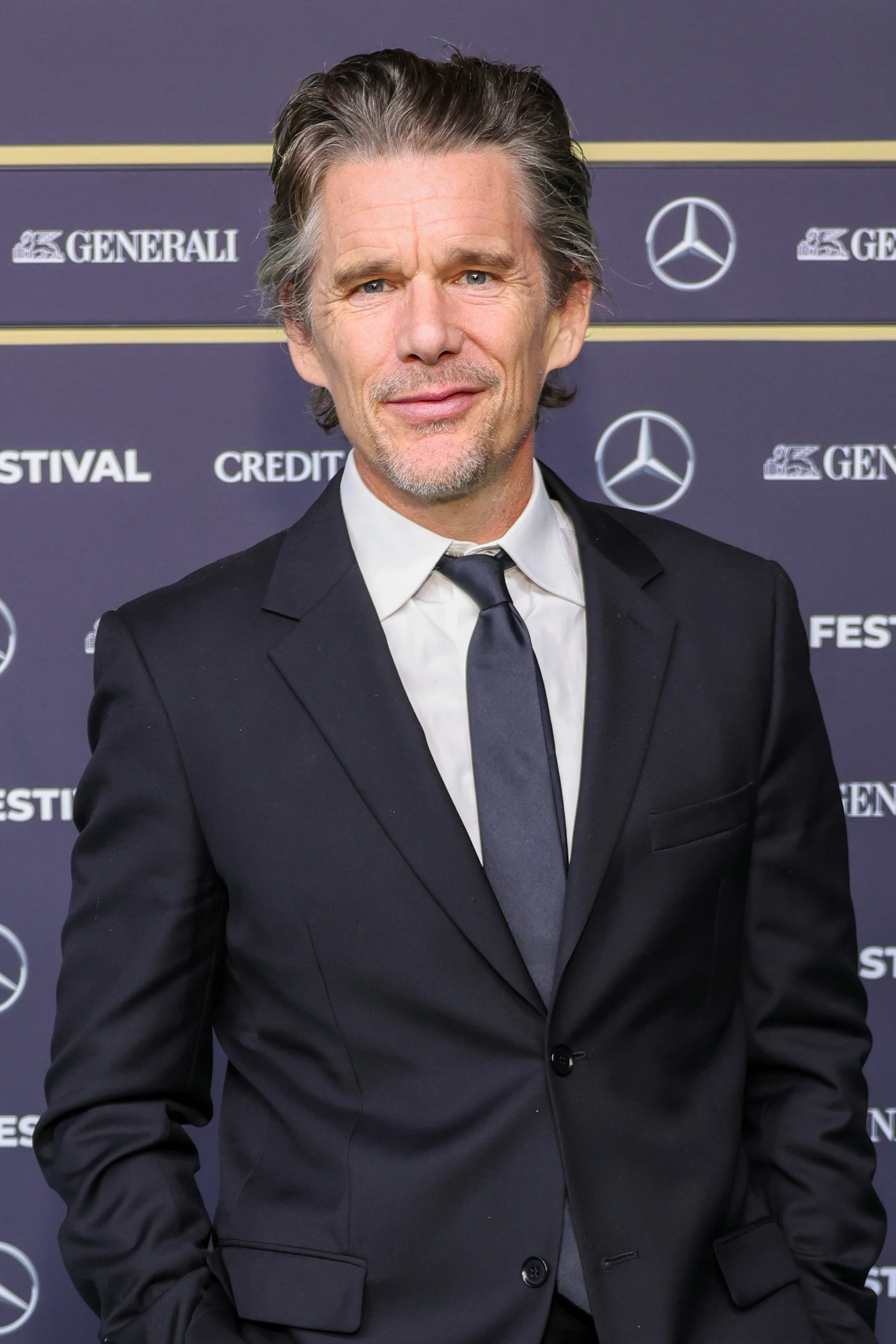 Male actor in a black suit, white shirt, and black tie at a formal event with a dark backdrop featuring logos.