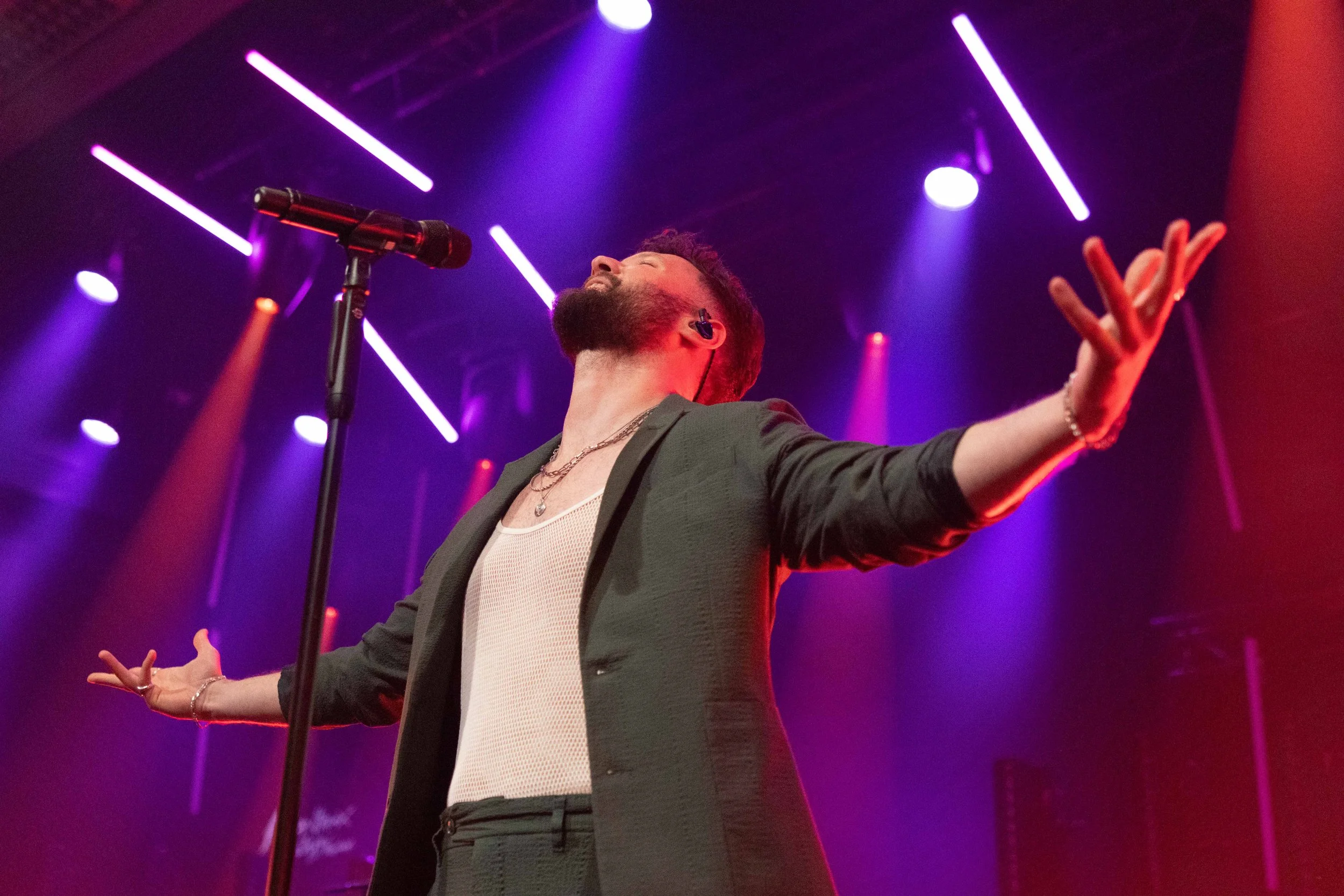 A male singer with a beard passionately performing on stage under colorful purple and red stage lights, with arms outstretched and eyes closed.