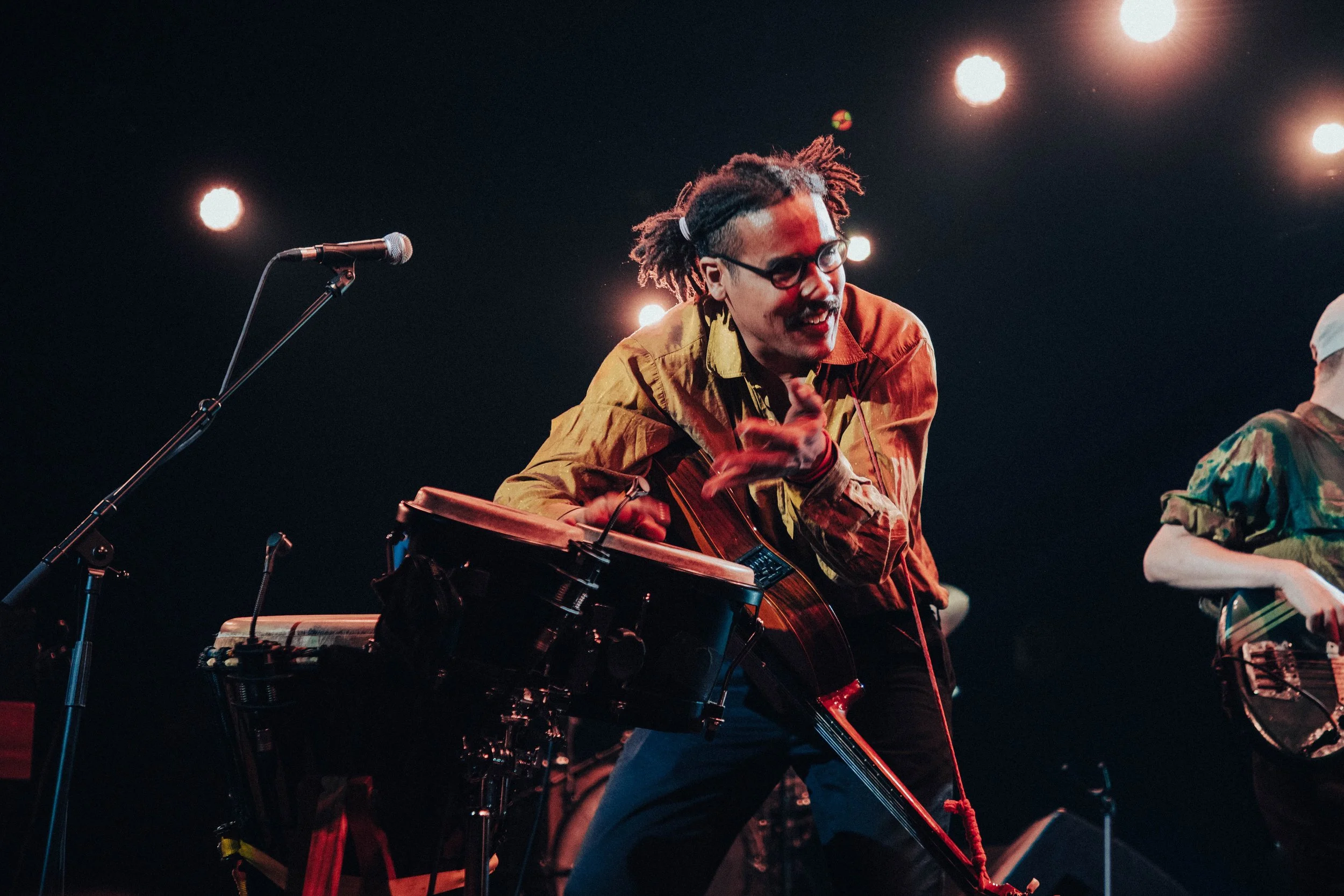 Performer with glasses and dreadlocks playing a guitar on stage under bright spotlights.