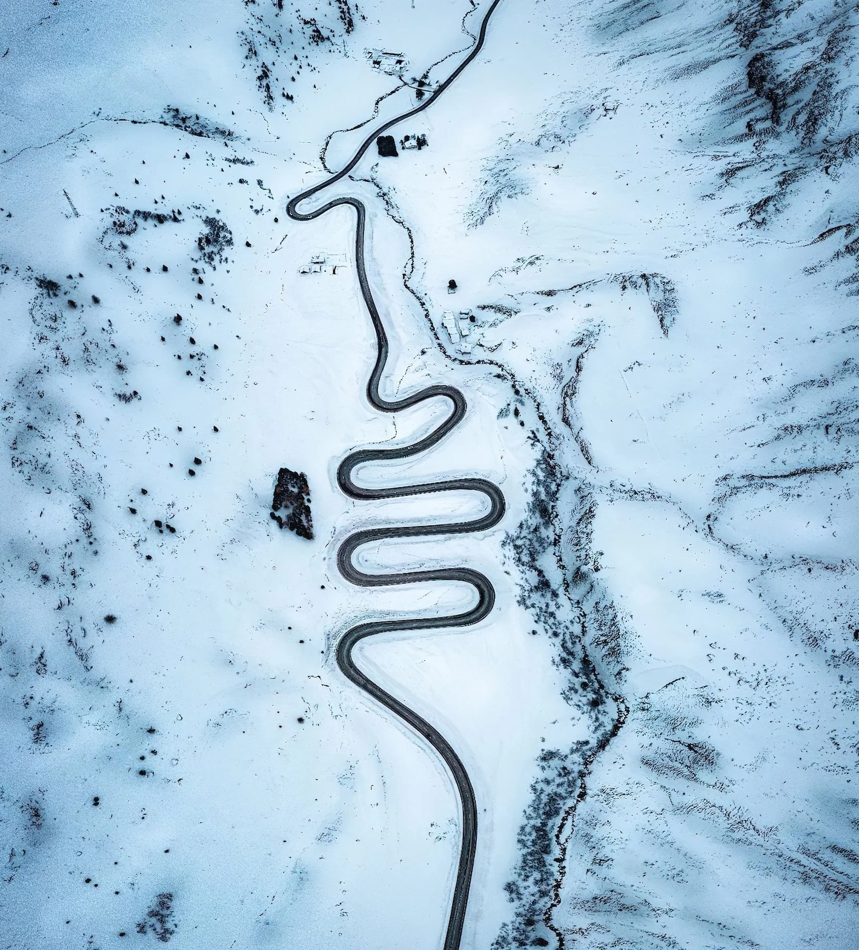 A winding mountain road covered in snow, seen from above, with sparse trees and buildings nearby.