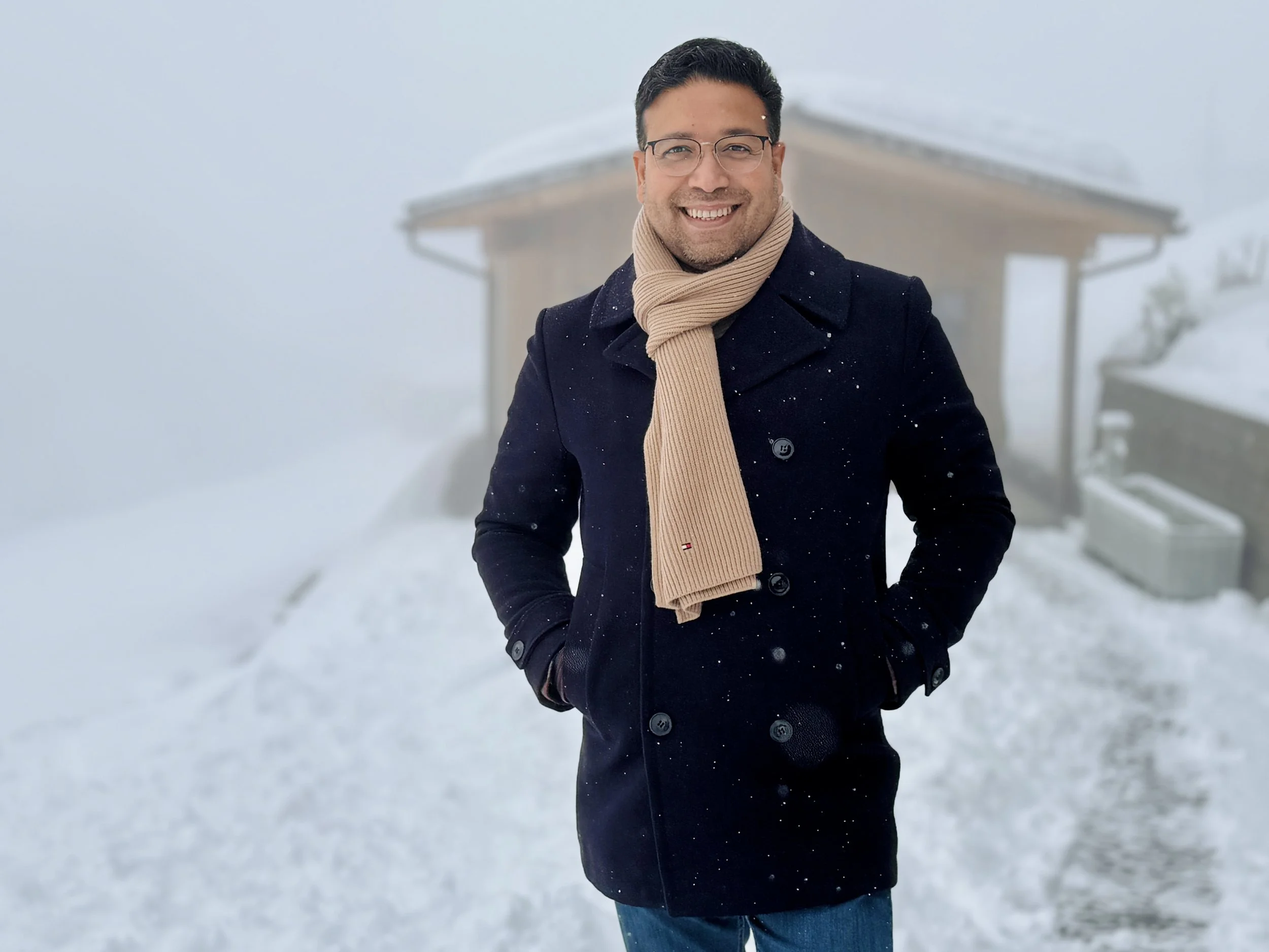 A man smiling outdoors in snowy weather, wearing glasses, a dark coat, and a beige scarf, with a wooden house and snow-covered ground in the background.