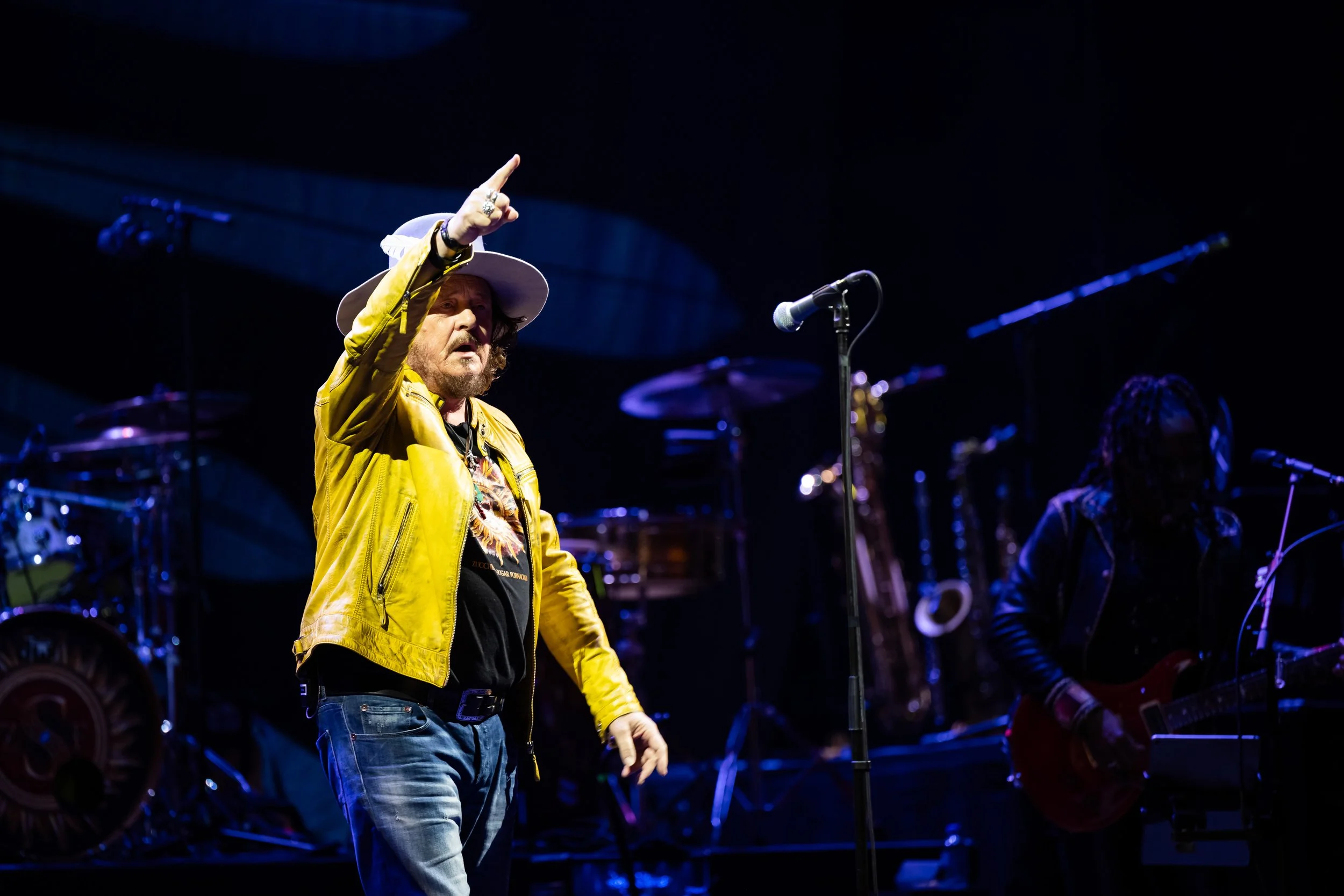 Male singer wearing a yellow jacket and wide-brimmed hat performing on stage with band instruments in the background.