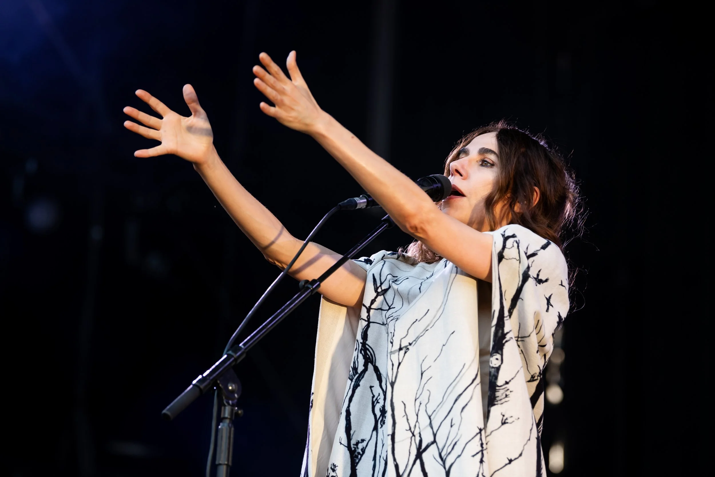 A woman singing into a microphone on stage, with her arms raised, against a dark background.