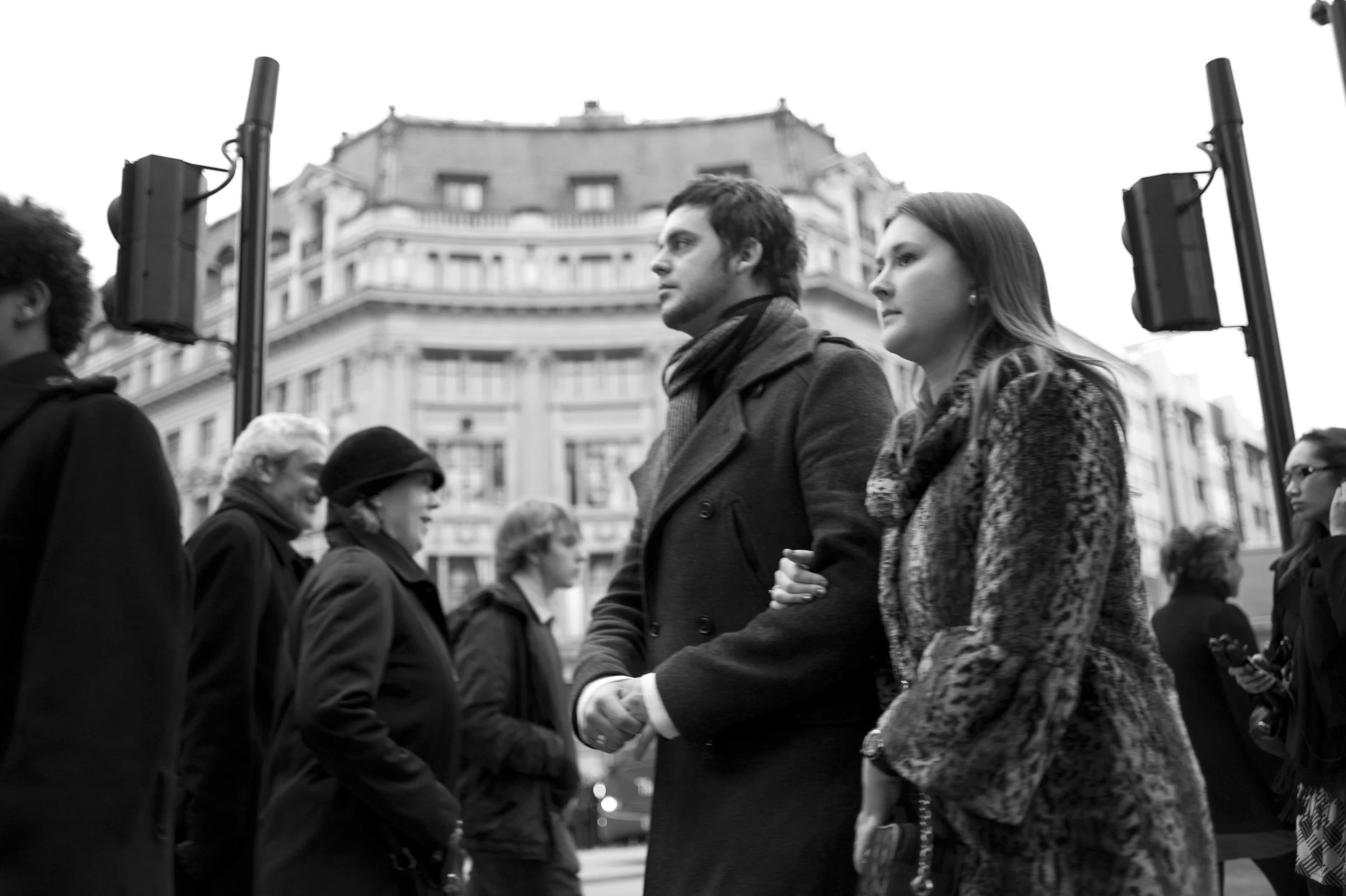 Black and white photo of a couple holding hands and walking on a city street, surrounded by other pedestrians, with a large historic building in the background.