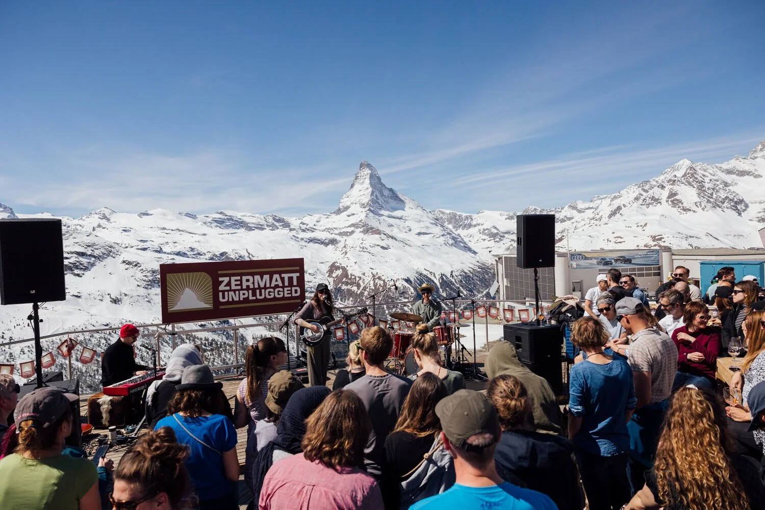 An outdoor music performance in the Swiss Alps with snow-covered mountains and the Matterhorn in the background. A band is playing on a stage labeled 'Zermatt Unplugged,' with an audience watching on a clear day.