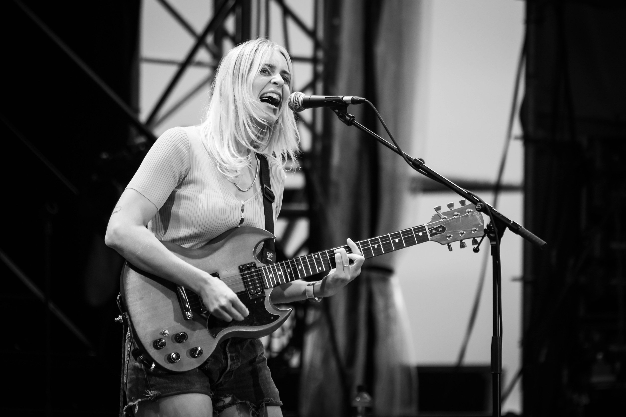 A woman with shoulder-length blond hair singing into a microphone while playing an electric guitar on stage