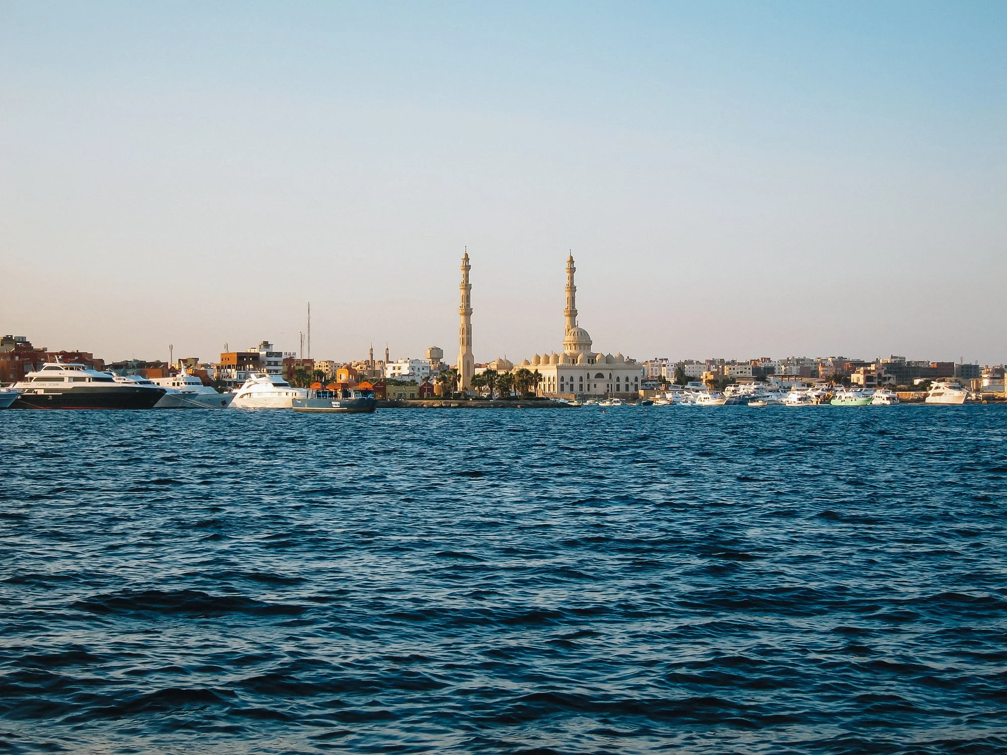 A view of a city shoreline with a mosque featuring two tall minarets, with boats docked in the water in the foreground.