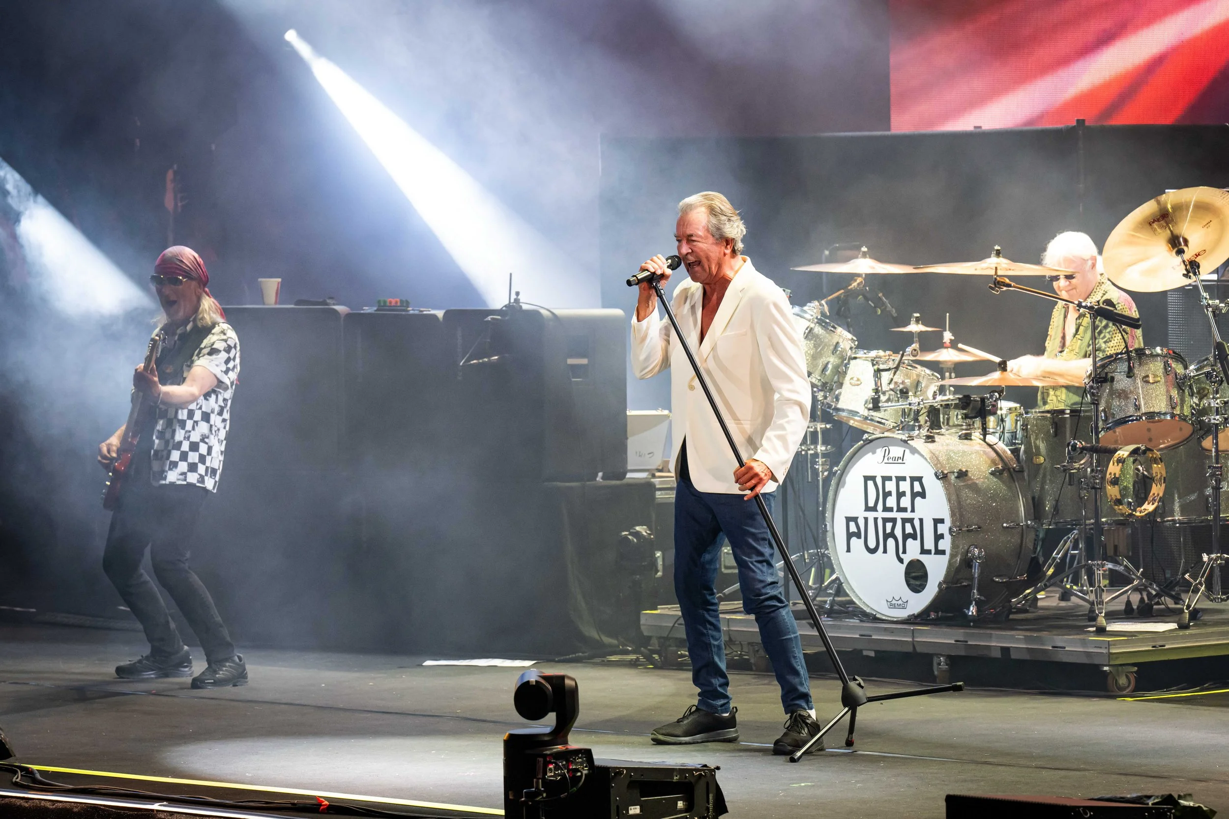 Band performing on stage with a vocalist in a white blazer, drummer, and guitarist, with 'Deep Purple' logo on the drum set, stage lights and smoke effects.