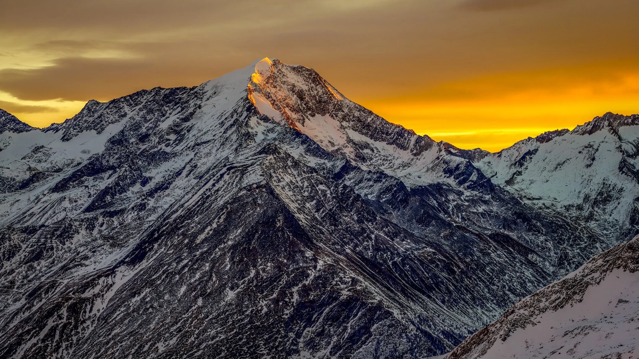 Snow-covered mountain peaks at sunset with an orange and yellow sky.
