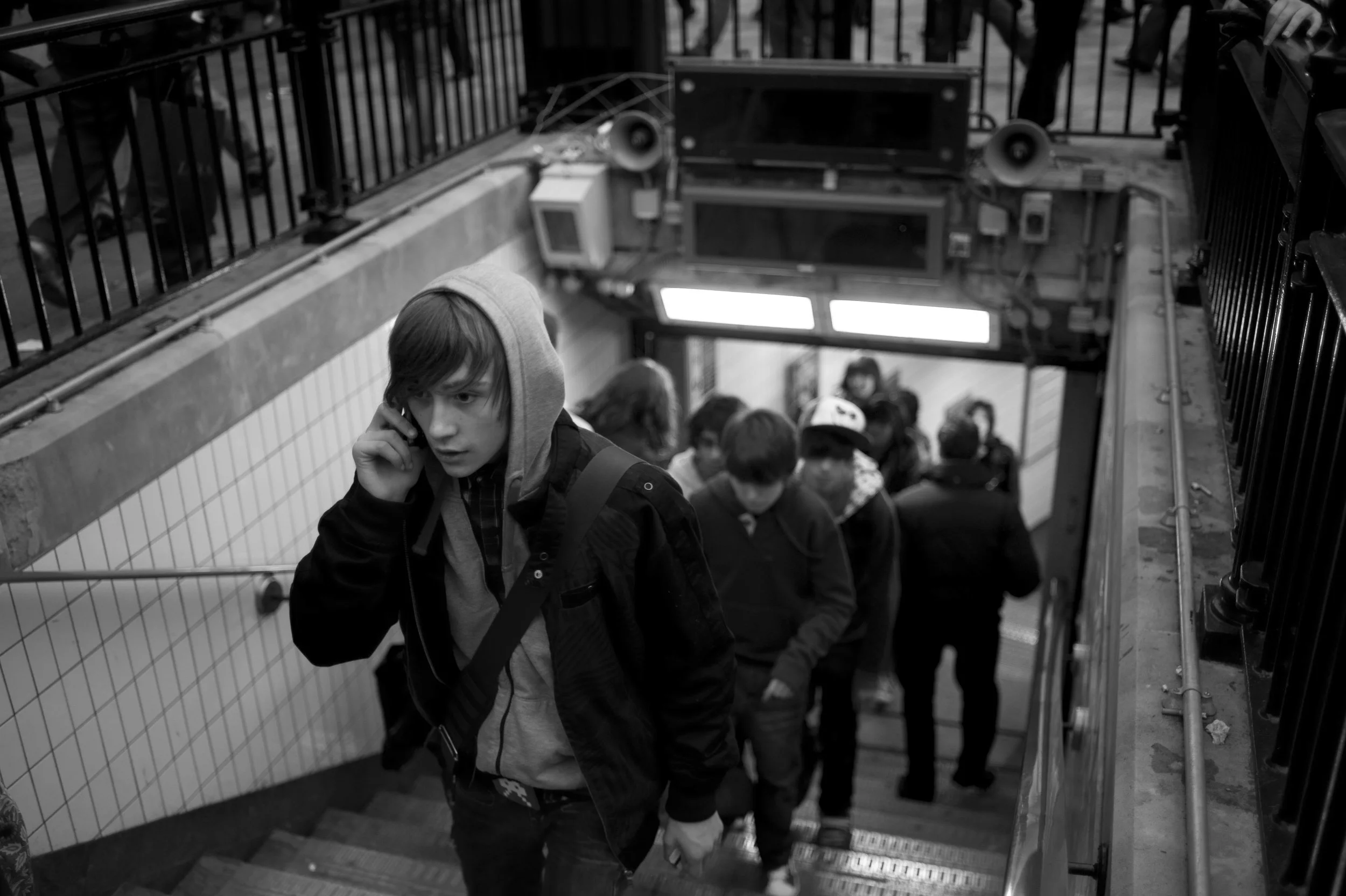 A young man with a hoodie talking on his phone while walking up the stairs in a subway station, surrounded by other commuters.