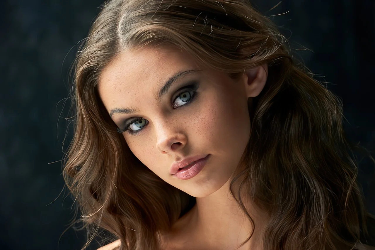 Close-up of a young woman with wavy brown hair, fair skin with freckles, and blue eyes, looking directly at the camera.