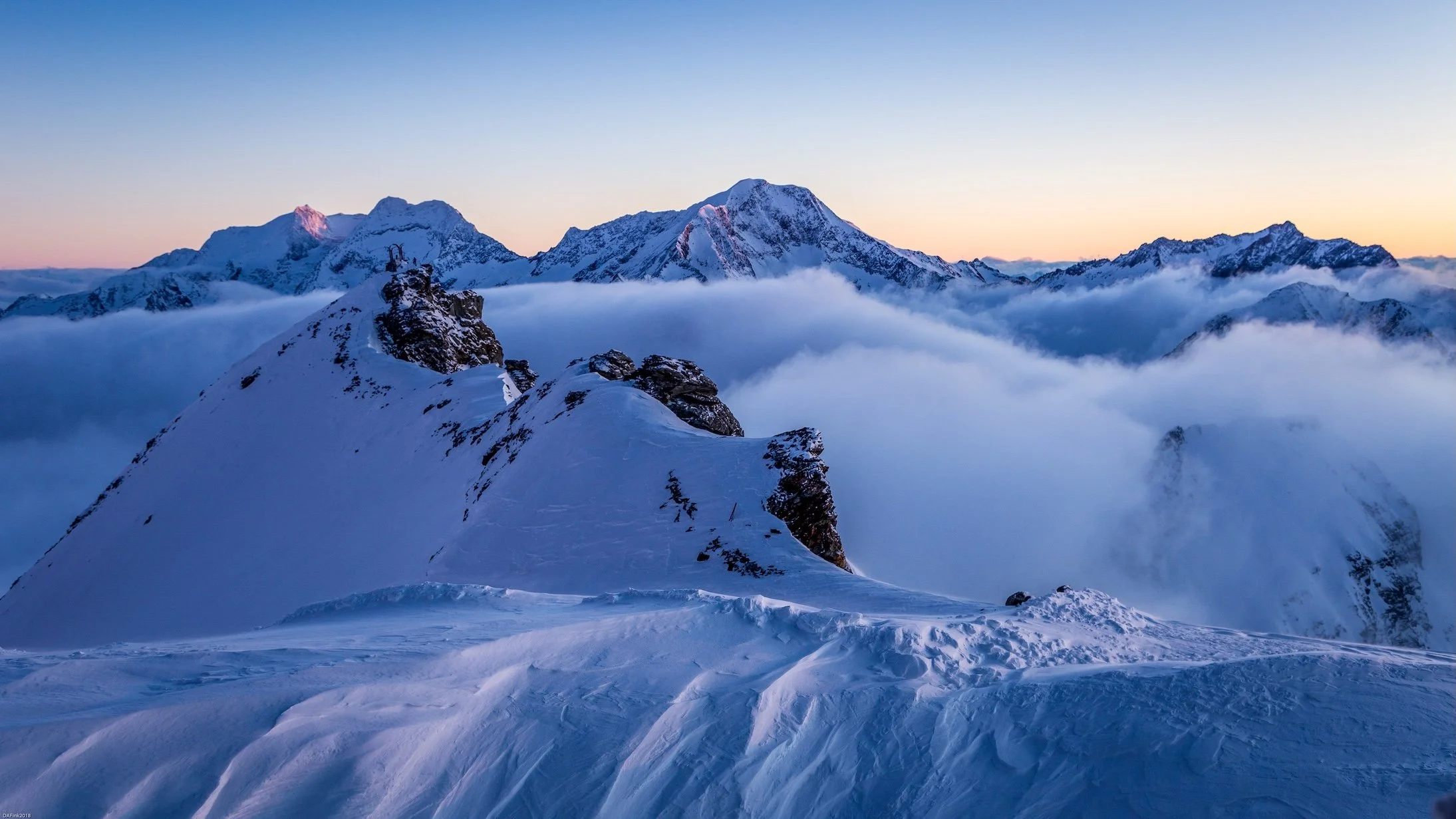 Snow-covered mountain range at sunrise with clouds below and a clear sky.