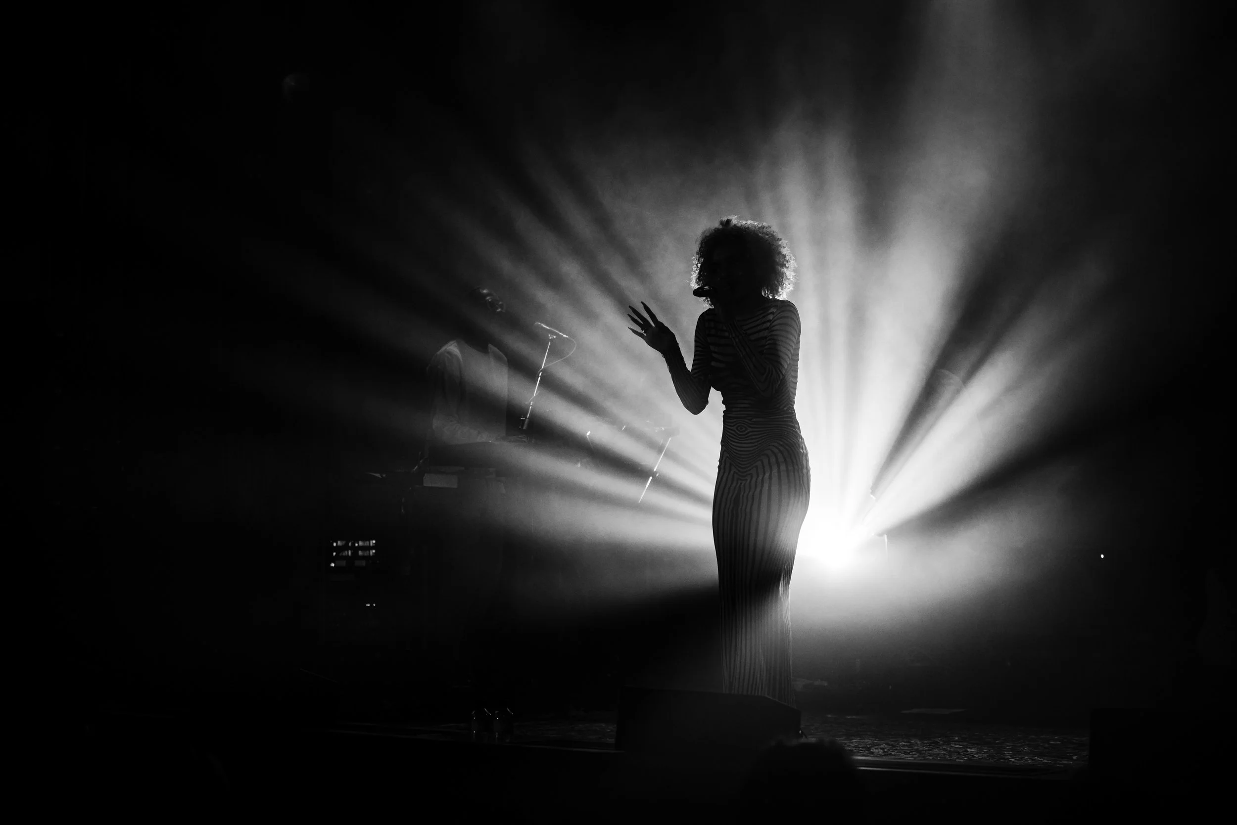 Silhouette of a singer on stage with dramatic backlighting creating rays of light radiating outward, accompanied by a keyboard or synthesizer player in the background.