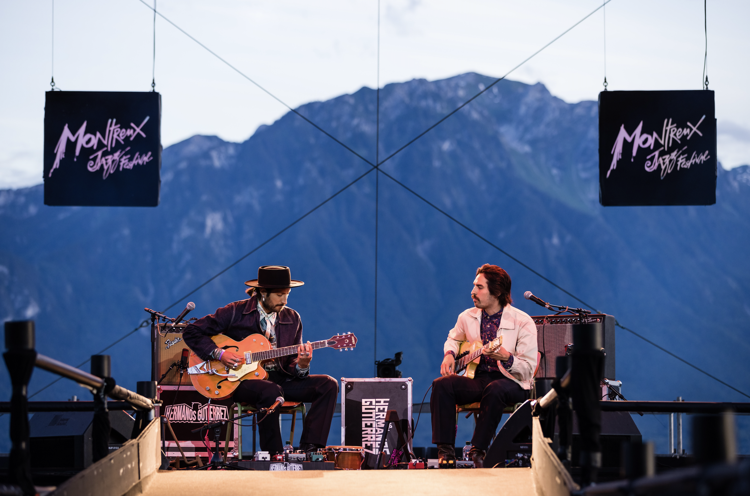 Two musicians playing guitars on an outdoor stage with mountain backdrop, banners reading Montreux Jazz Festival, and equipment around them.