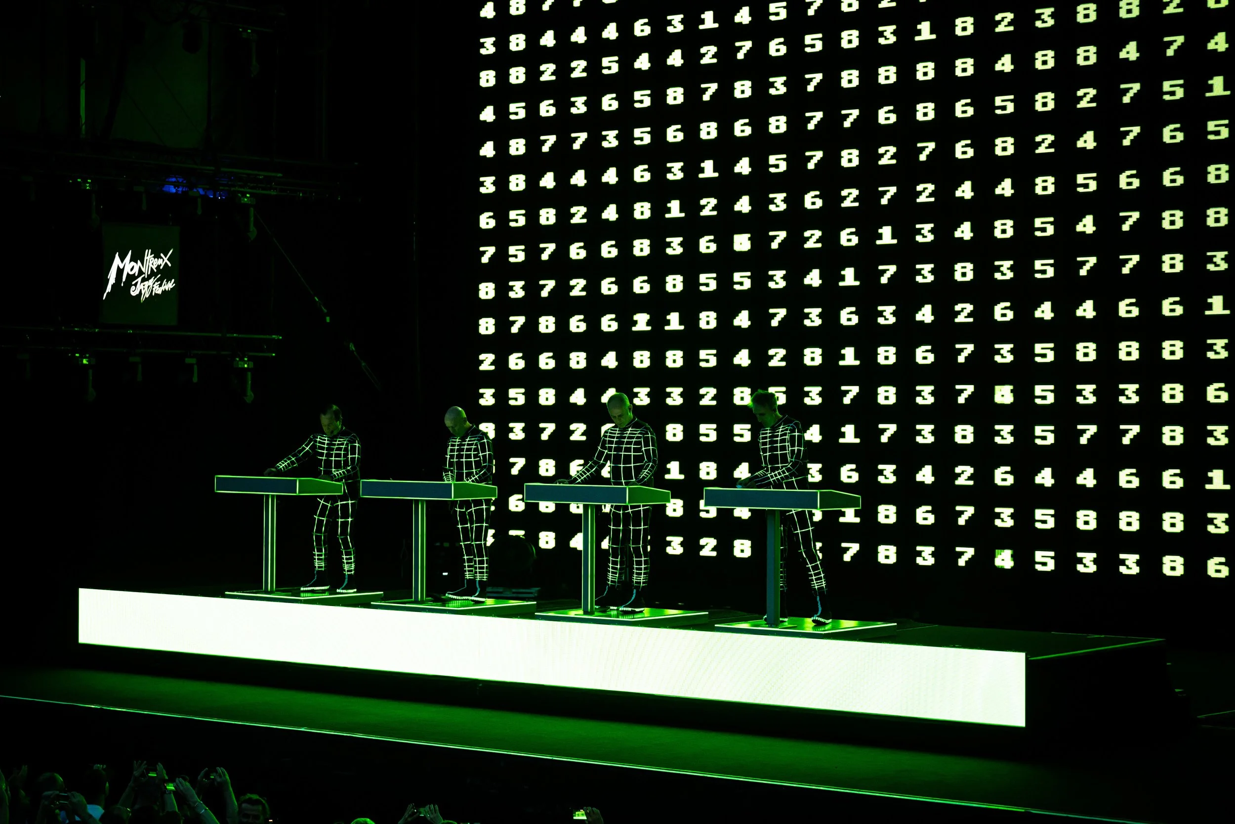 Four performers in black and white checkered outfits stand at multi-colored electronic keyboards on a stage with a large screen showing green numbers in the background.