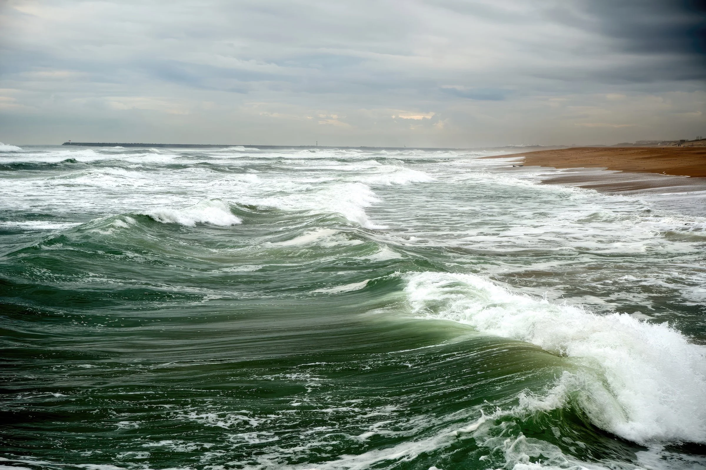 Overcast sky with dark clouds over a beach with green waves crashing onto the shore and a sandy coastline in the distance.