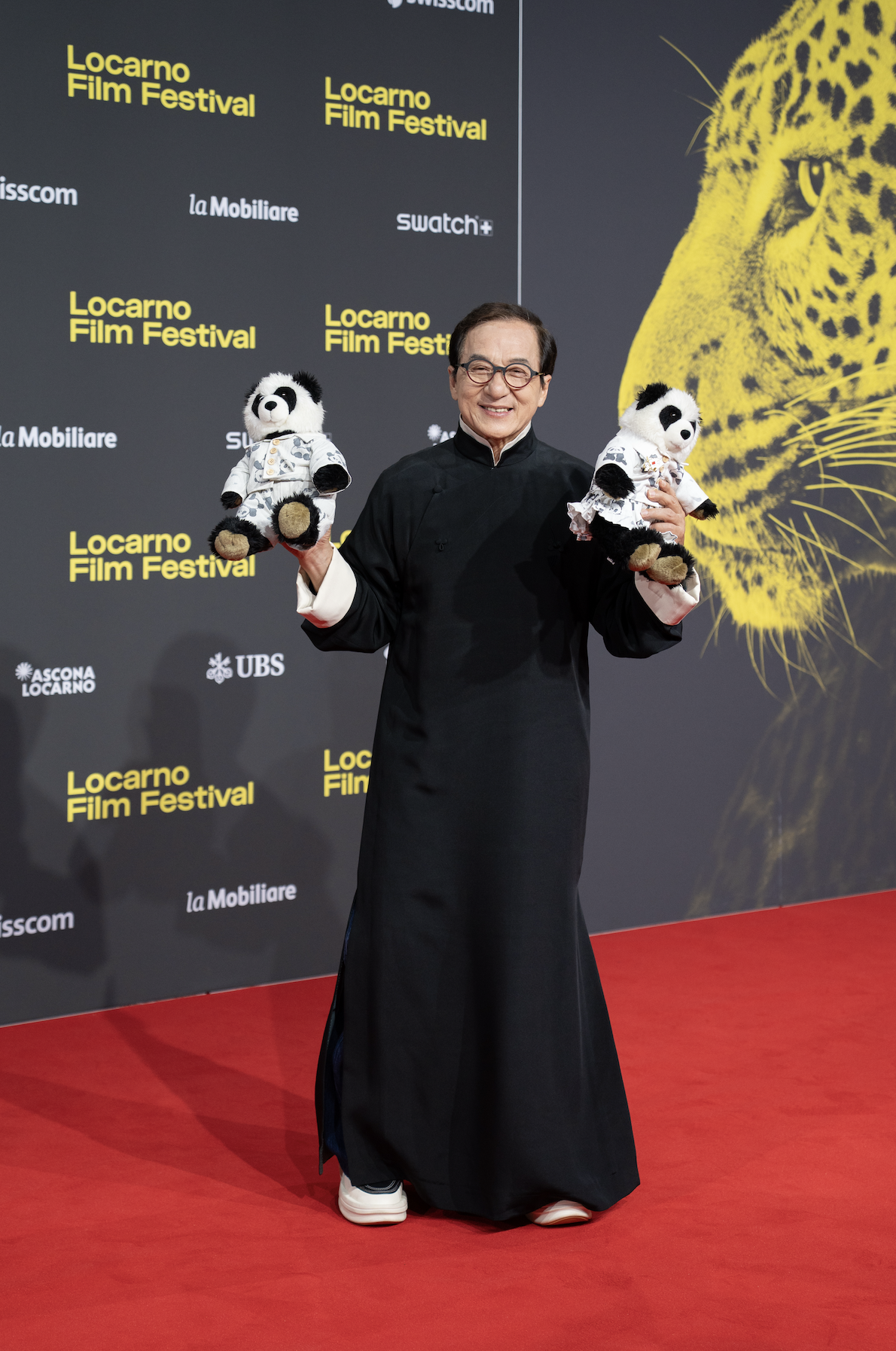 A man wearing glasses and traditional black attire smiling and holding two panda plush toys dressed in clothing at the Locarno Film Festival, with a backdrop featuring the festival's name and event sponsors and a large image of a yellow leopard.