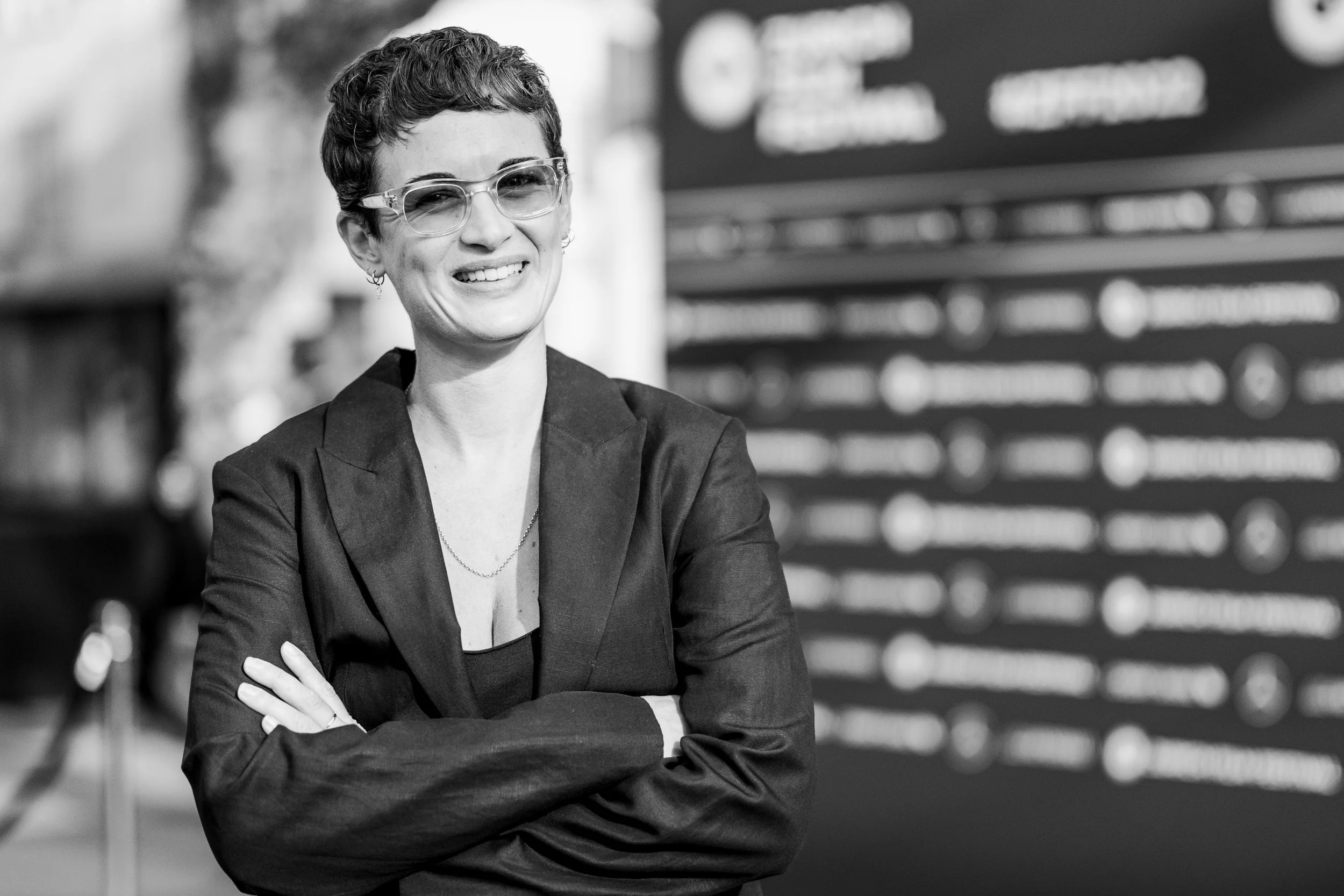 A woman with short curly hair, wearing glasses and a blazer, smiling with her arms crossed in front of a blurred background.