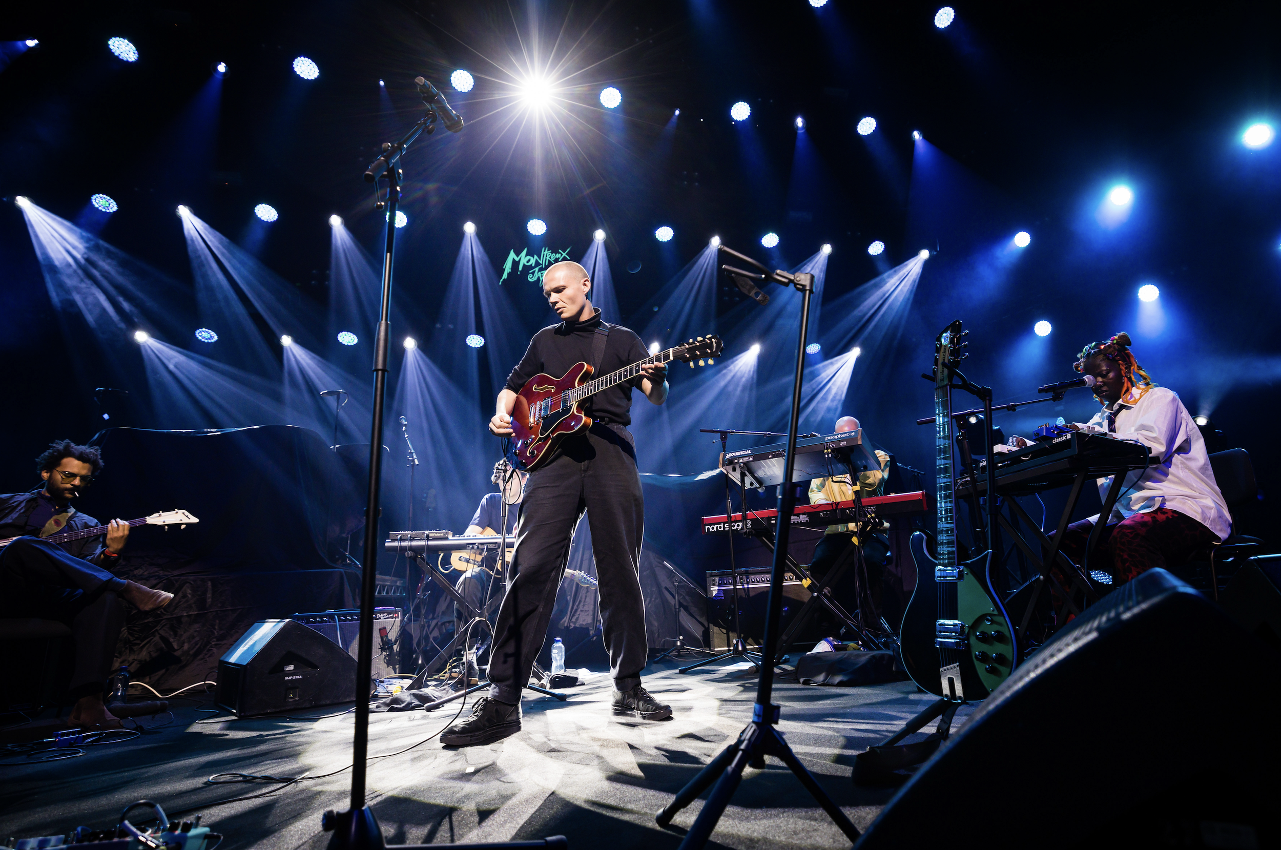 A band performing live on stage with multiple musicians playing guitars, keyboards, and drums, under blue stage lights at the Montreux Jazz Festival.
