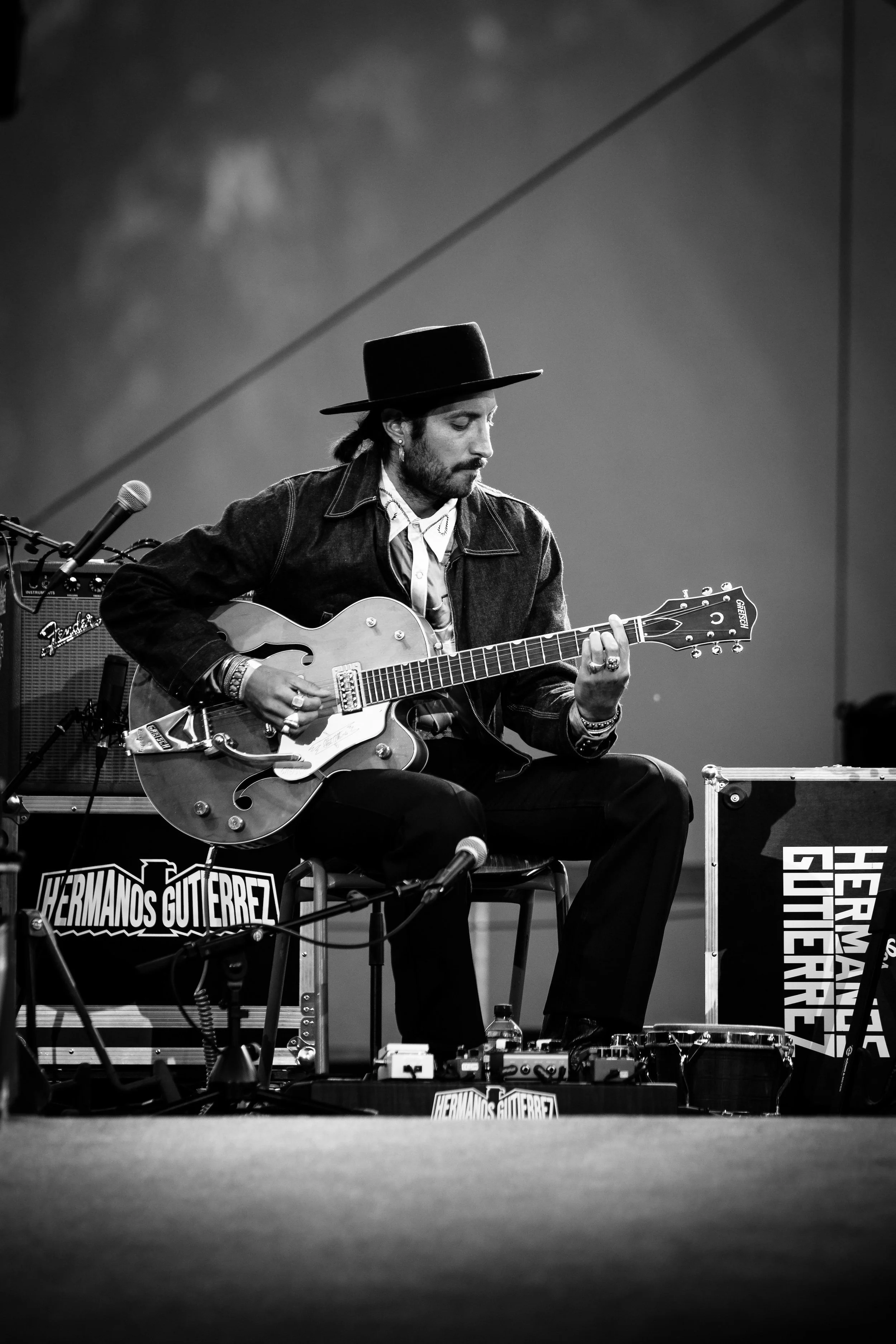 Black and white photo of a man playing an electric guitar, sitting on a chair with a hat and denim jacket, on a stage with musical equipment and a banner reading 'Hermanos Guiterrez'.