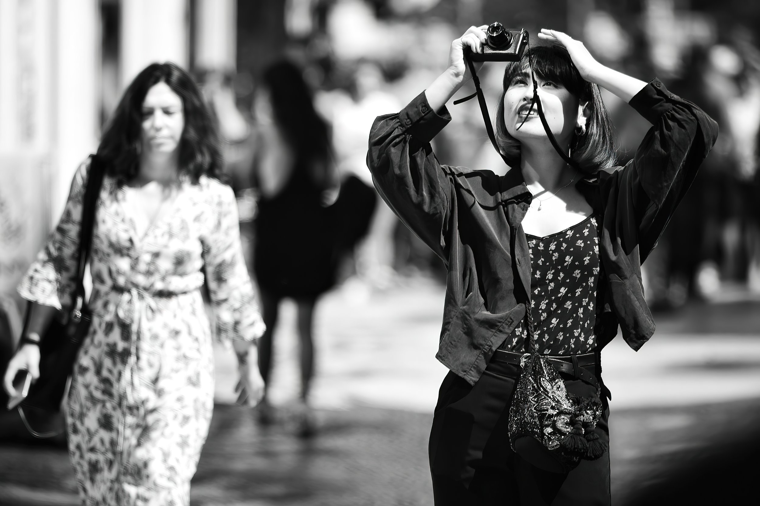 A young woman takes a photo with a camera on a busy city street. She has shoulder-length hair and is wearing a floral top and a dark jacket. Other pedestrians walk in the background.
