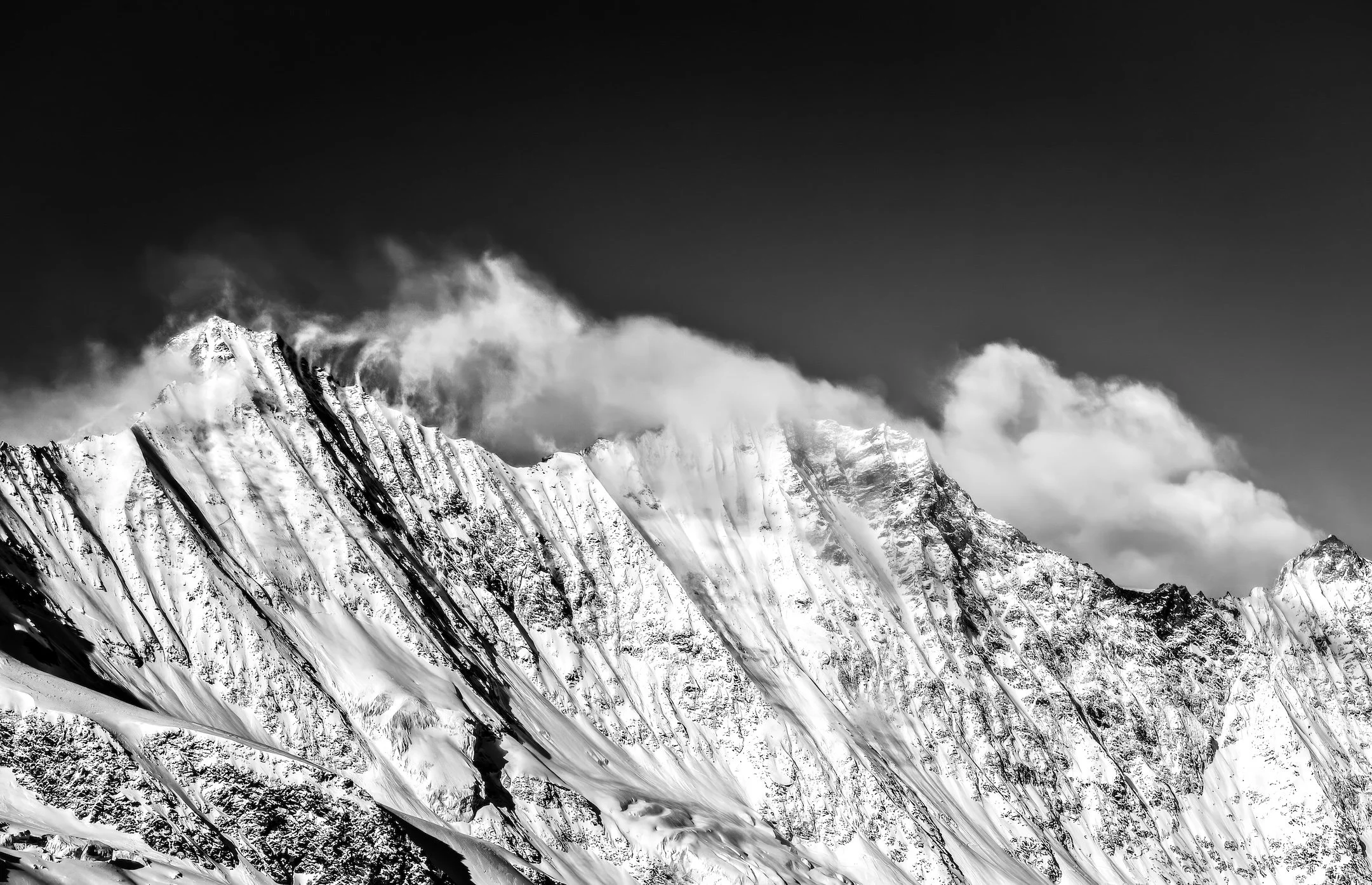Snow-covered mountain range with steep ridges and clouds above, in black and white.