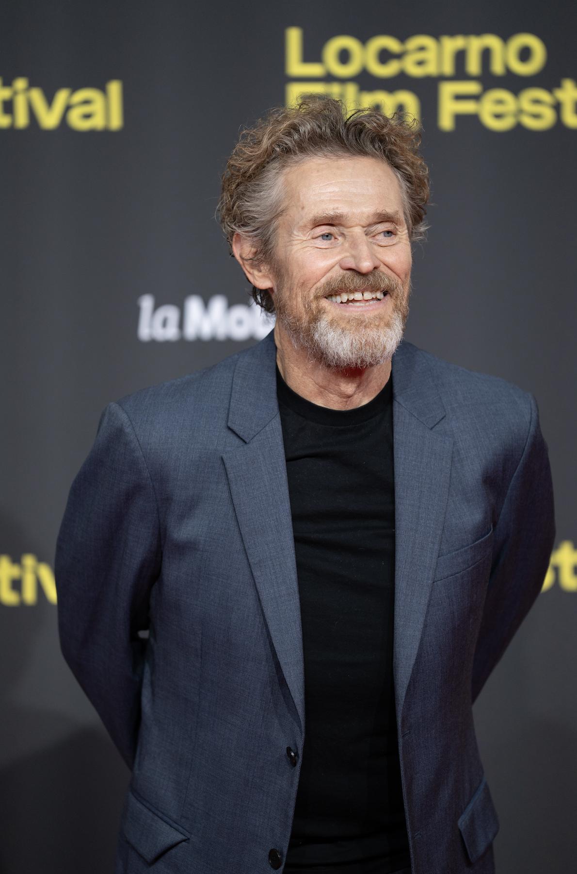 A man with a beard and curly hair, wearing a dark blue blazer and a black shirt, smiling at a film festival event in front of a black backdrop with yellow and white text.