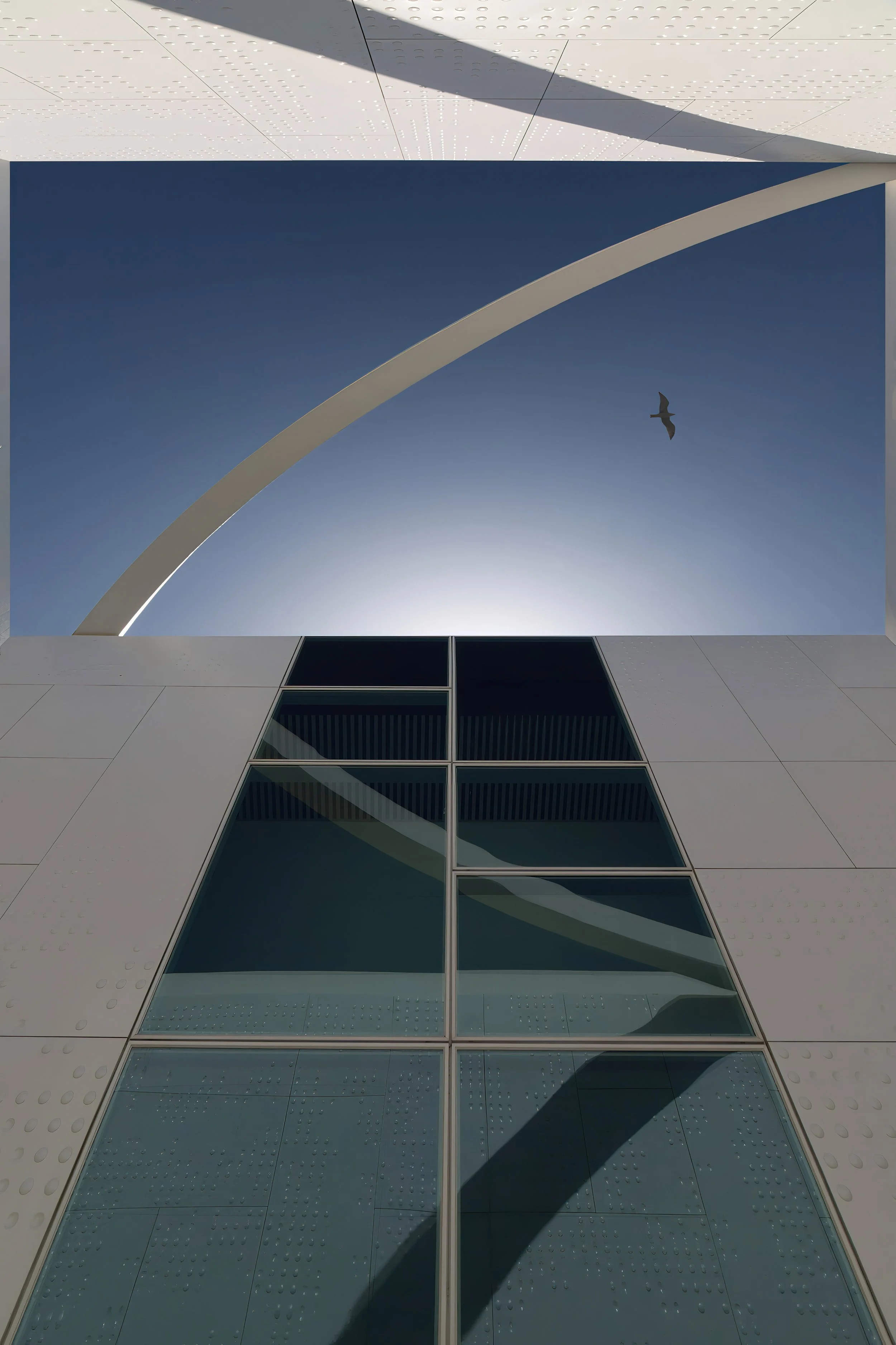 Looking up through a building's opening at a clear blue sky with a bird flying overhead and the arches of the building framing the sky.