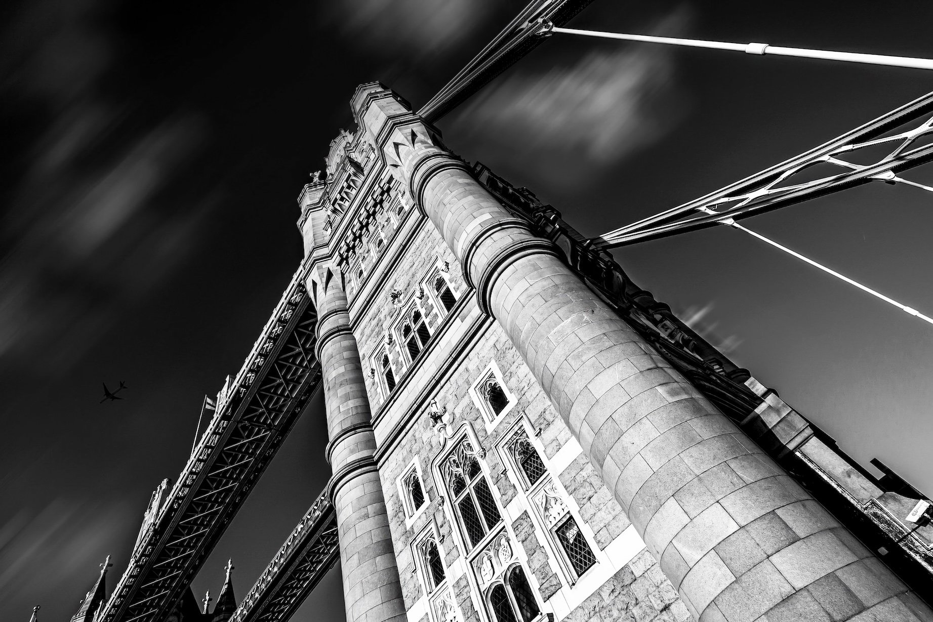 Black and white photo of Tower Bridge in London, seen from below with the bridge's towers and suspension cables visible, sky with clouds, and an airplane in the background.