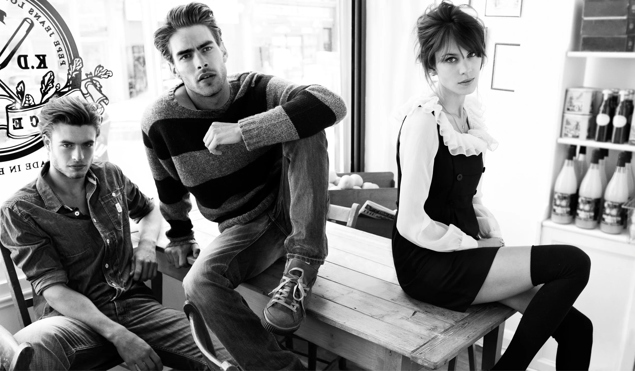 Three young adults in a cafe, two men and one woman, sitting on a wooden table with a store display in the background.