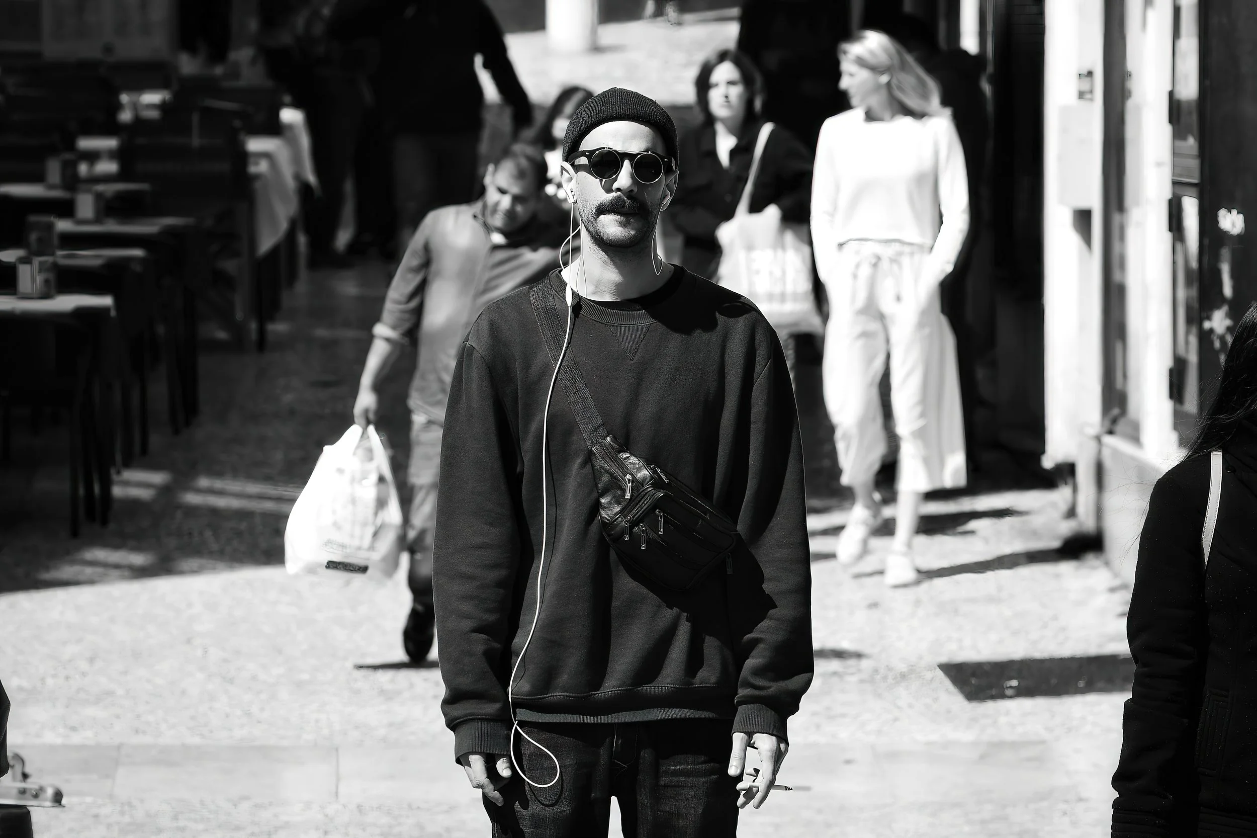 A man wearing sunglasses, a beanie, and a cross-body bag is standing on a busy city street, with other pedestrians walking in the background.