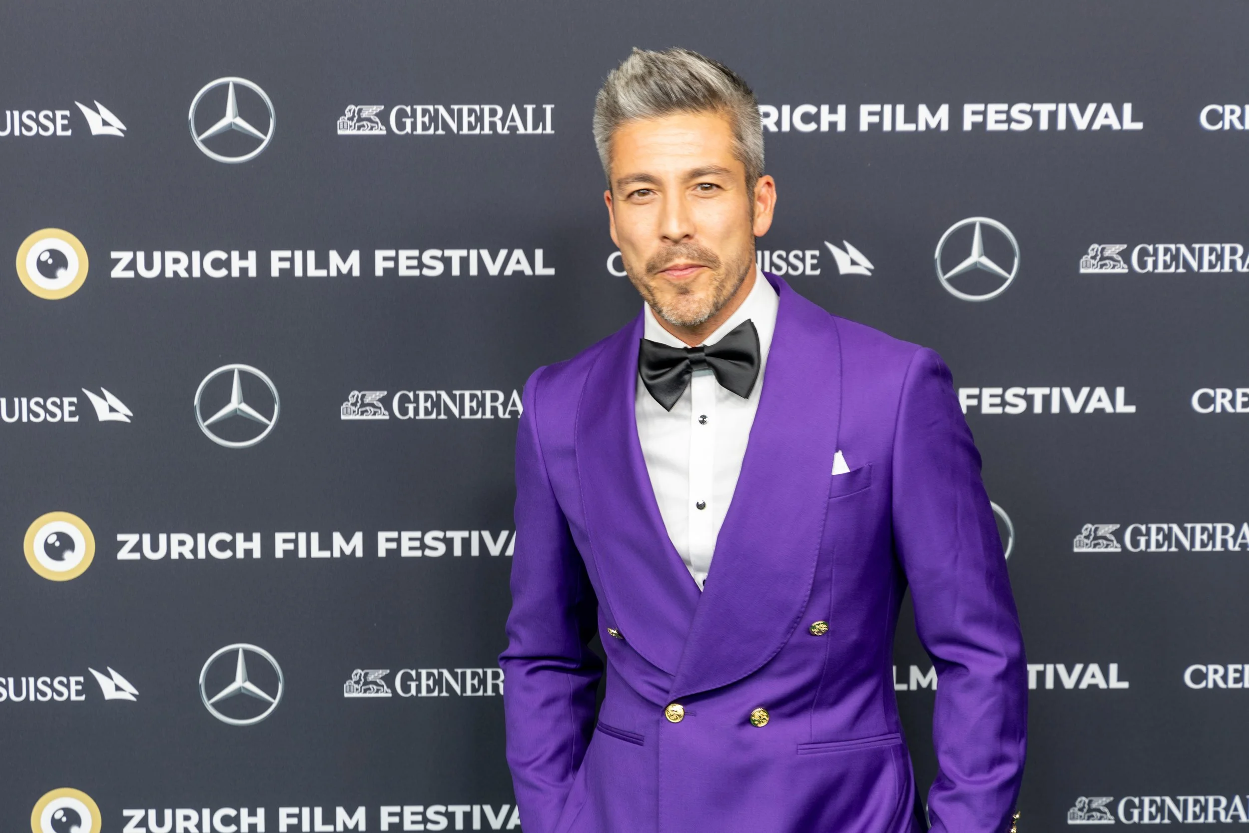 Man in a purple tuxedo with a black bow tie standing in front of a backdrop with 'Zurich Film Festival' logos.