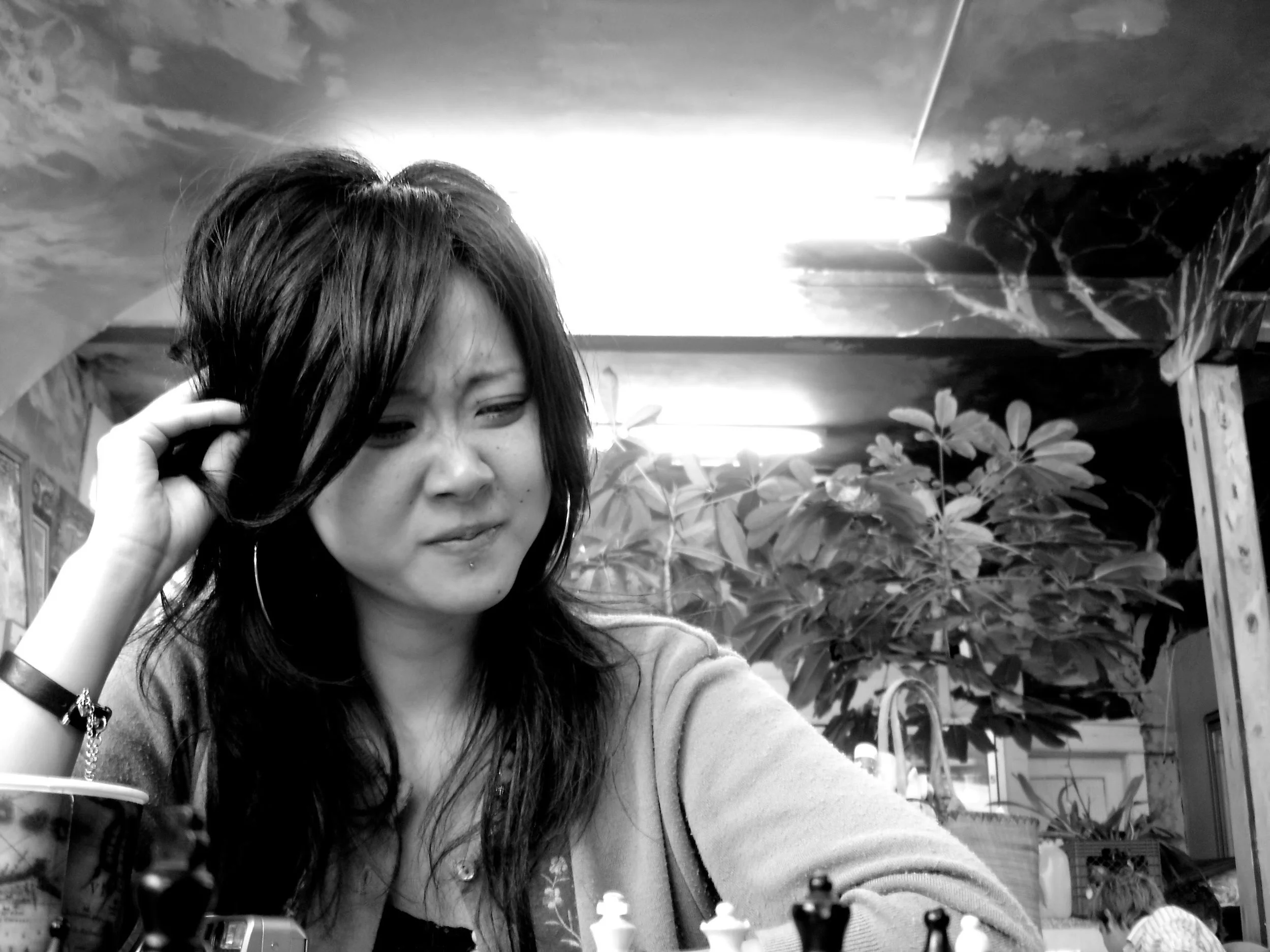 A woman with dark, wavy hair, wearing large hoop earrings, staring at a chessboard with a puzzled expression in a room with plants and bright lighting.