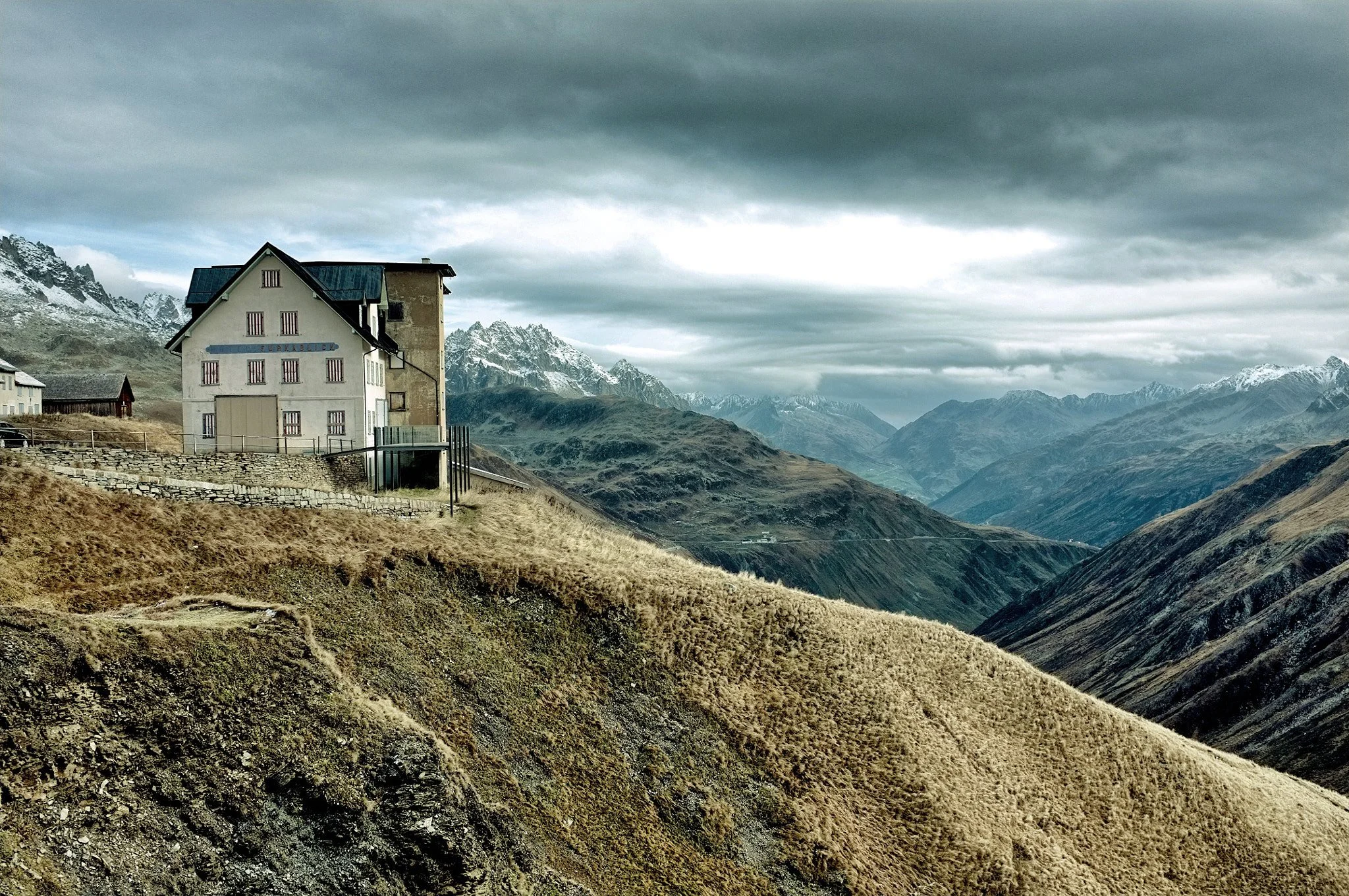 A house on a hill overlooking rugged mountain terrain with snow-capped peaks under a cloudy sky.