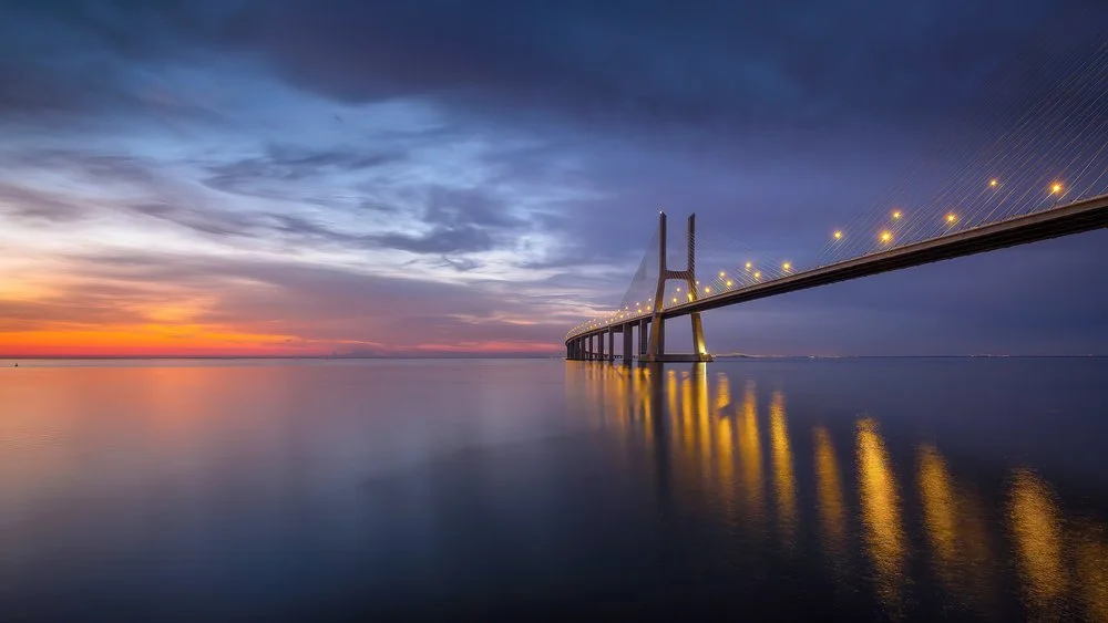 A large suspension bridge with illuminated cables over a body of water during sunset, with colorful sky and reflections on the water.