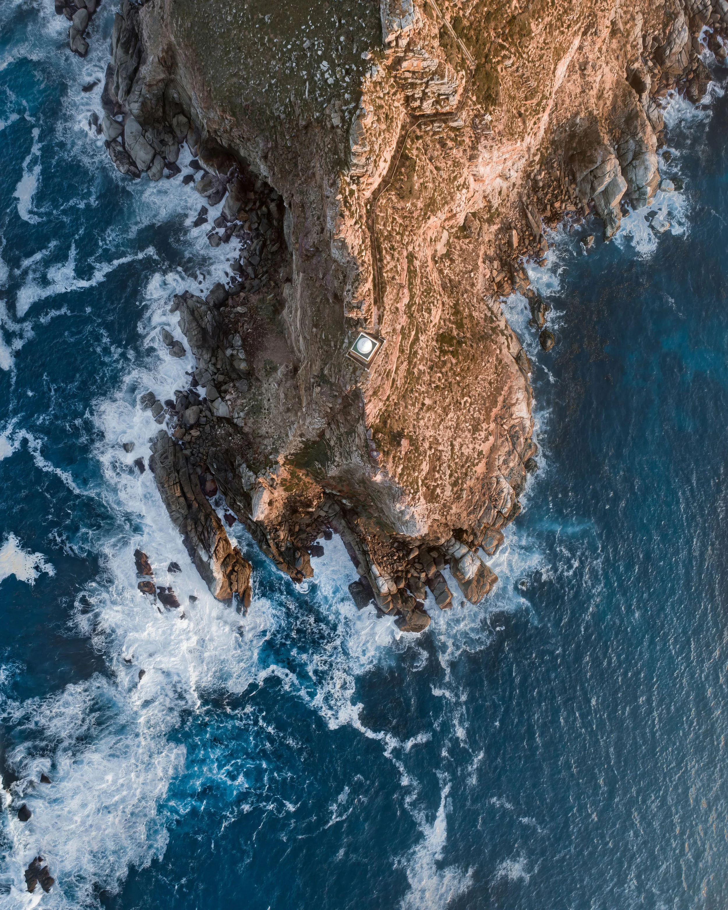 Aerial view of a rocky coastal cliff with waves crashing against the rocks and a small building or structure situated on the edge of the cliff.