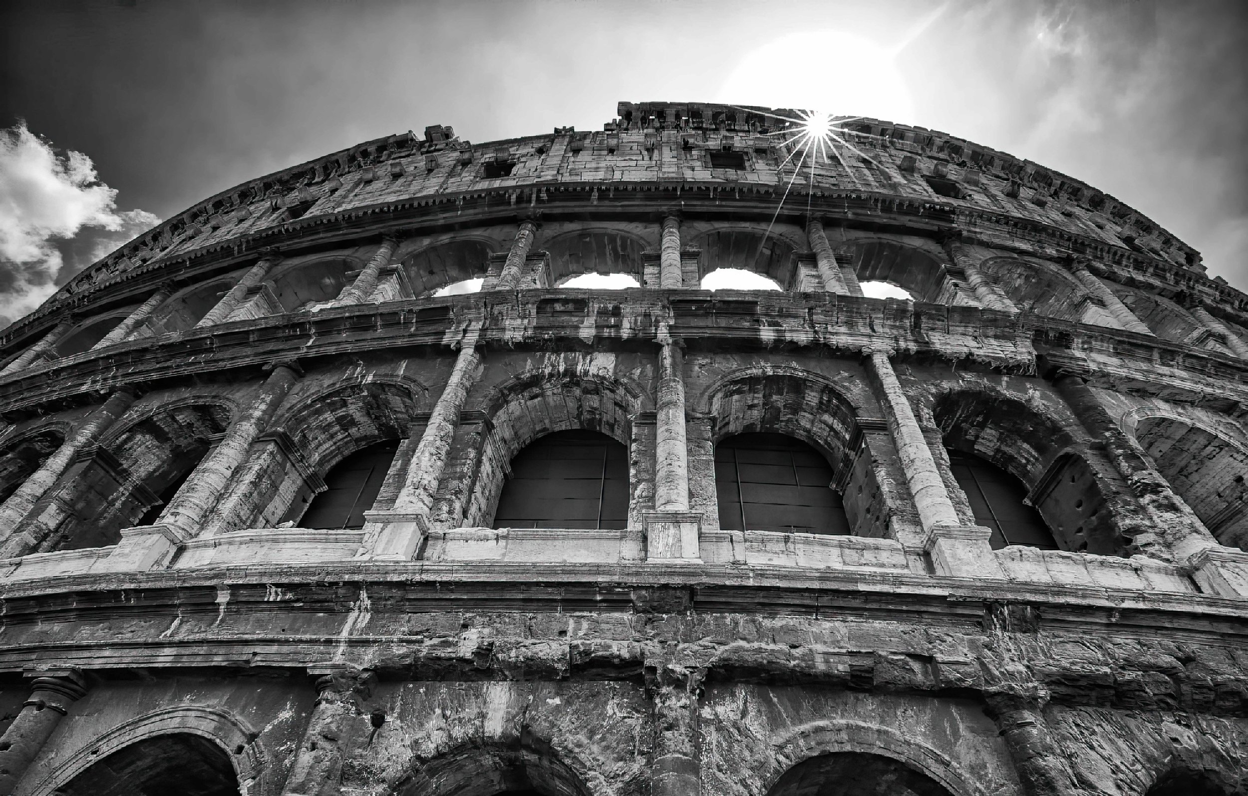 Black and white photo of the Colosseum from a low angle with the sun shining behind it.