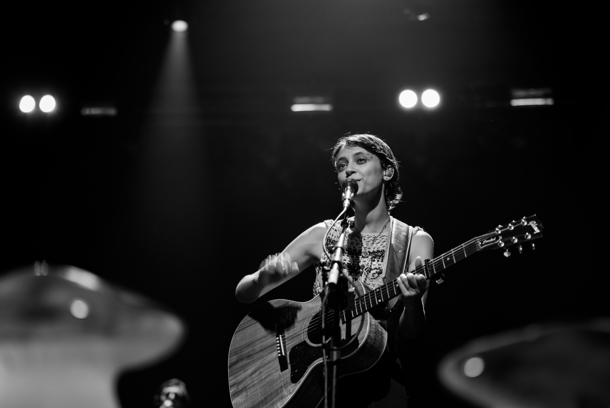 A young woman with short hair playing an acoustic guitar and singing into a microphone on stage, lit by spotlights in a black and white photo.