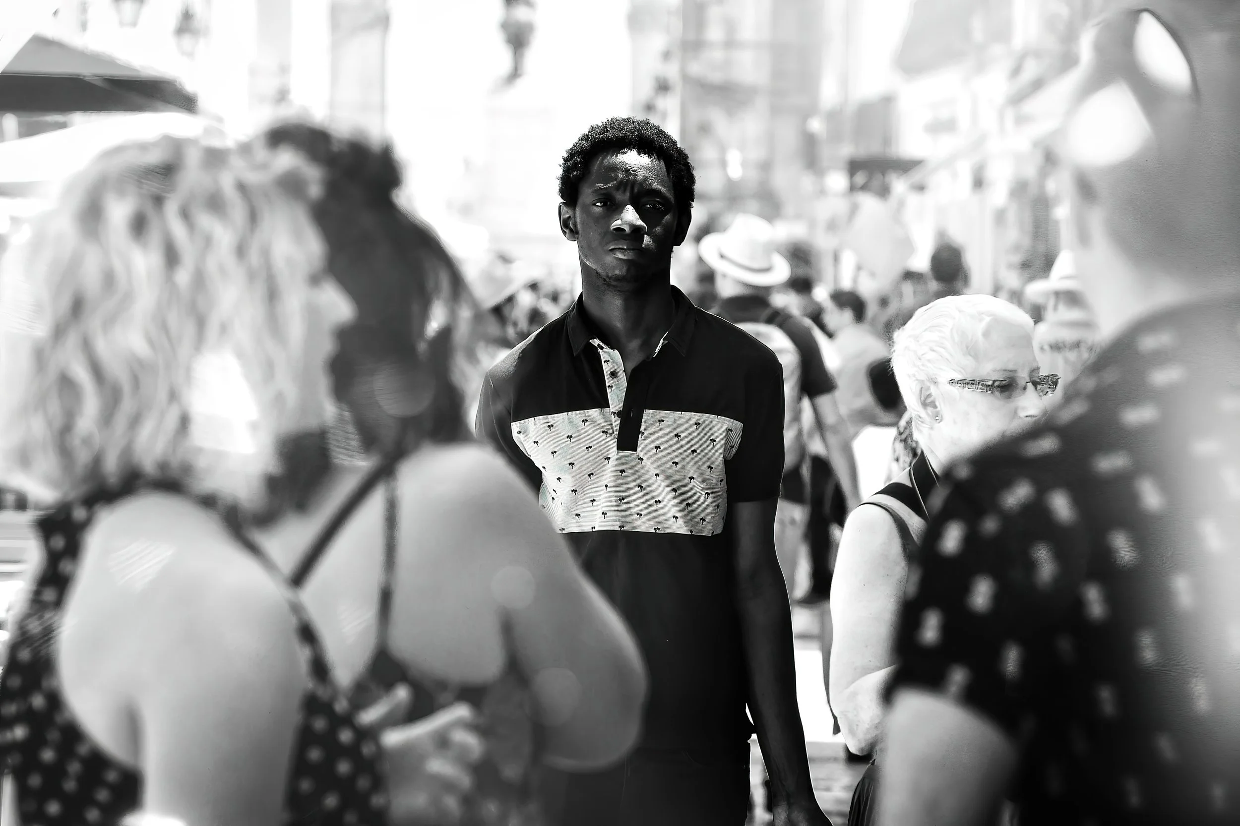 A young man with dark skin and curly hair looks directly at the camera amid a busy street scene with pedestrians, some wearing hats, in black and white.