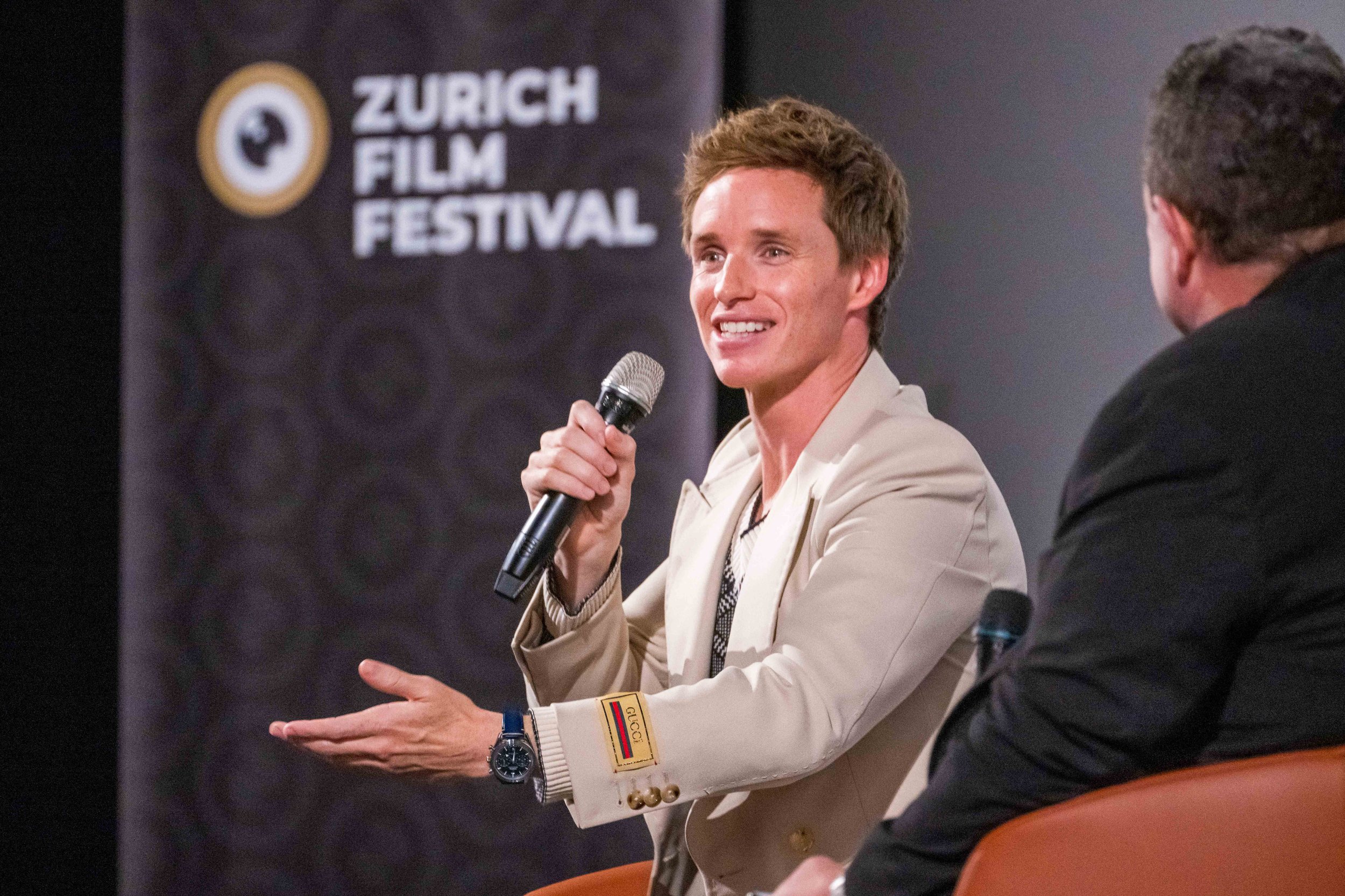 A man holding a microphone and speaking at the Zurich Film Festival. He is wearing a light-colored blazer with Gucci and flag patches on the sleeve, and a watch. There is another person partially visible sitting next to him.