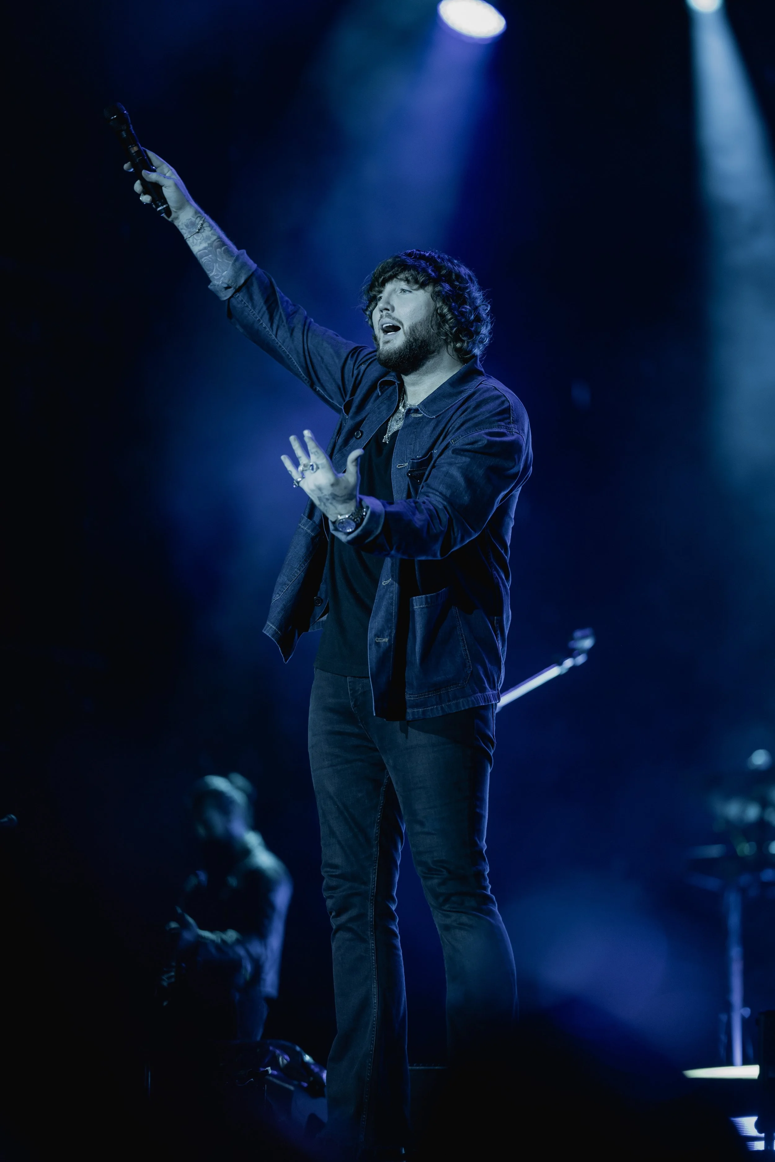 Male singer performing on stage with microphone, raising his hand, surrounded by stage lights and a dark background.