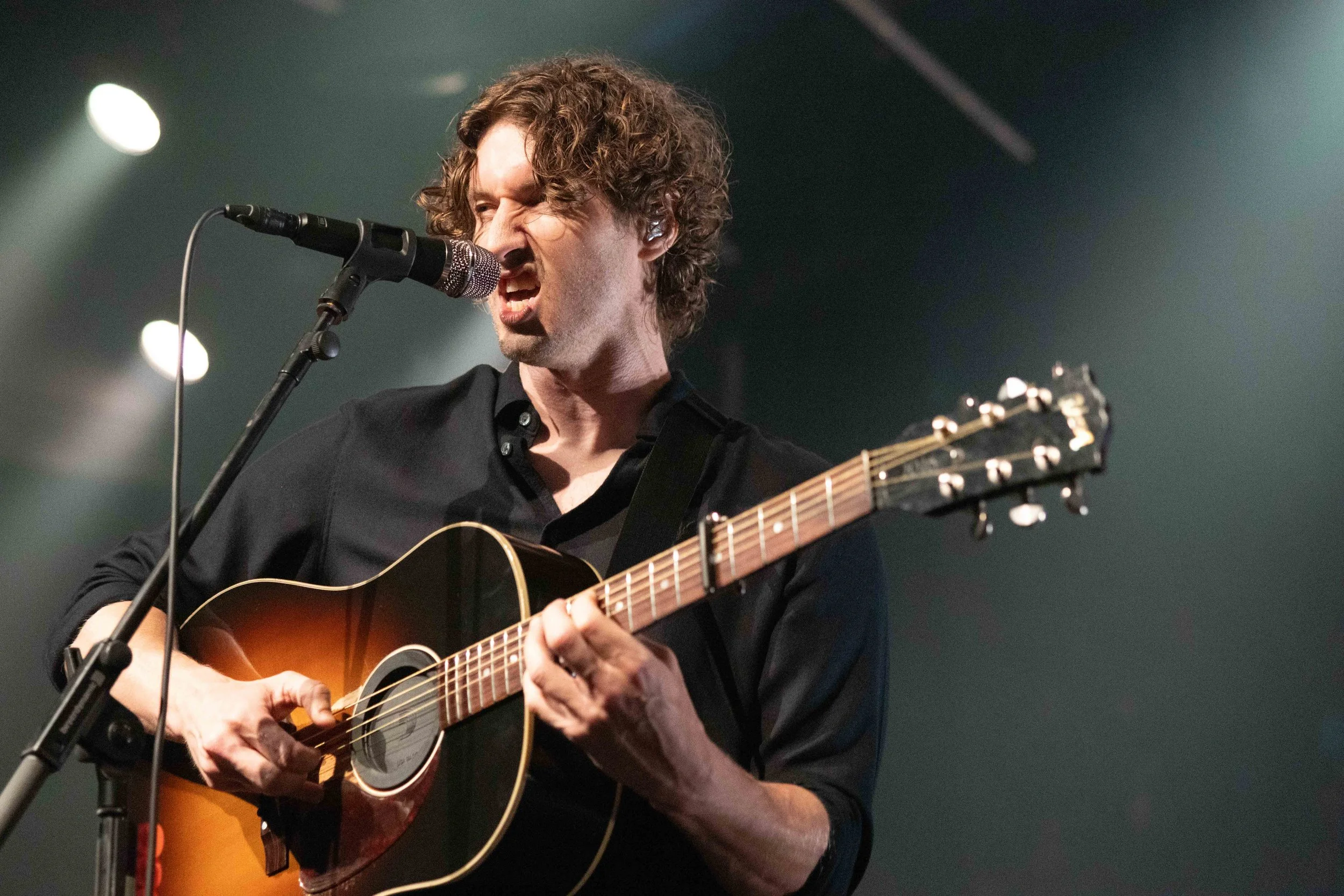 A man with curly hair sings into a microphone while playing an acoustic guitar on stage during a live performance.