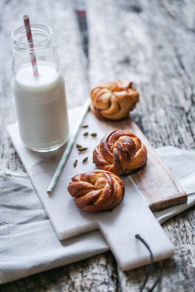 Three cinnamon rolls on a wooden and marble serving board, a glass bottle of milk with a striped straw, and some scattered pumpkin seeds, all on a rustic wooden table.