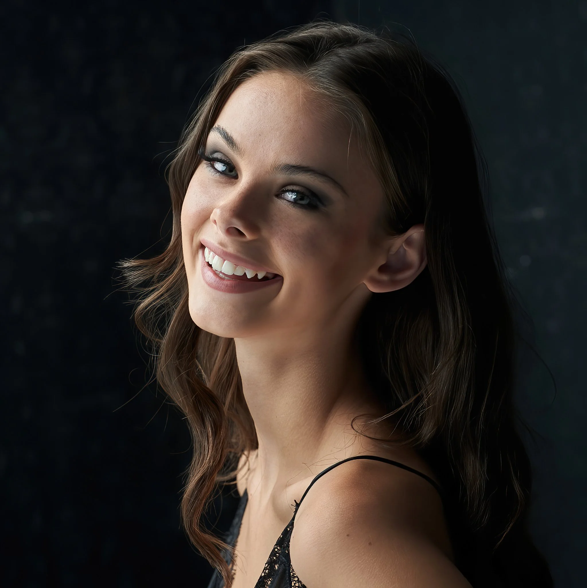 A smiling woman with wavy brown hair, blue eyes, and light makeup, wearing a black lace camisole against a dark background.