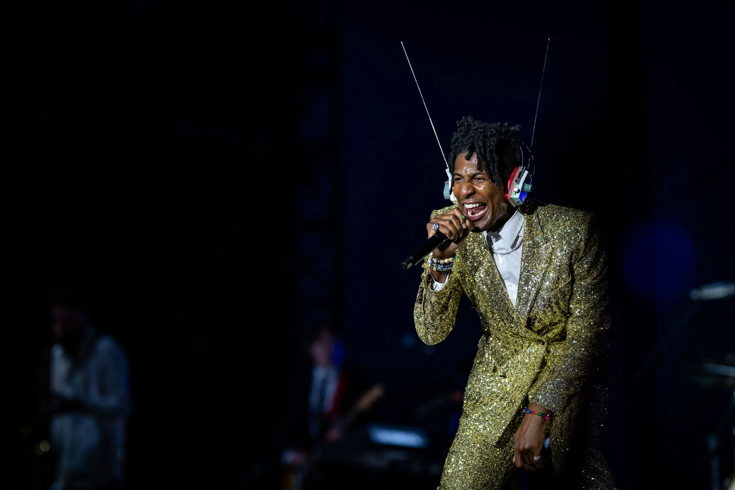 A male performer in a glittering gold suit singing into a microphone, wearing headphones with antennas, on a dark stage with other musicians in the background.
