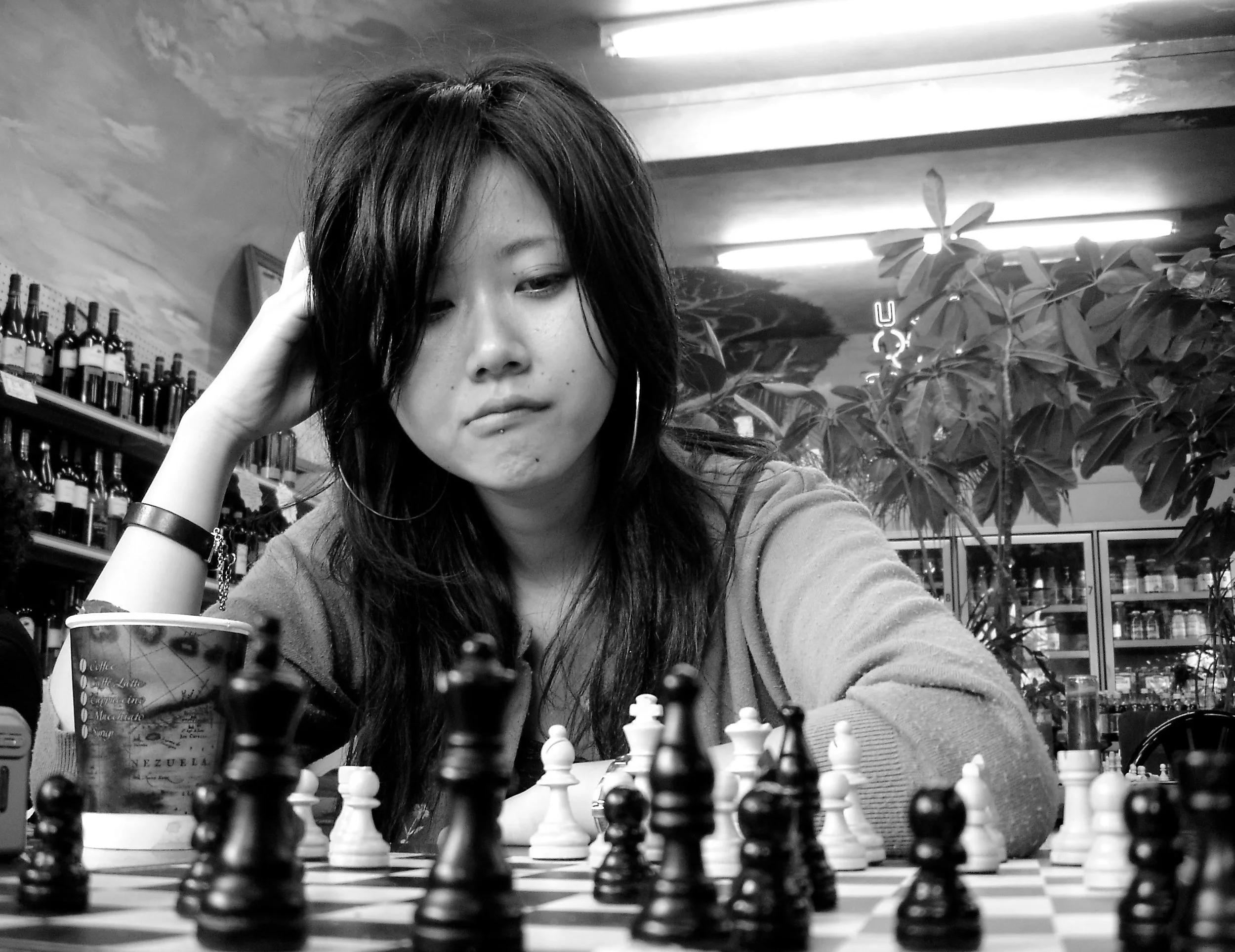 A woman with dark hair and large hoop earrings looks at a chessboard, resting her head on her hand. She is in a cafe or store setting with wine bottles on shelves behind her and a large leafy plant to her right.