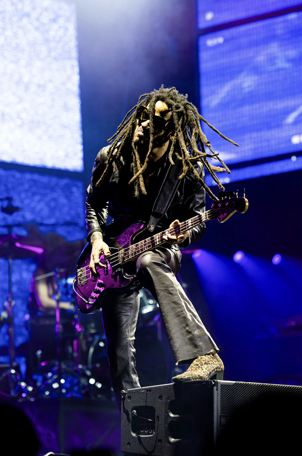 Musician with dreadlocks playing an electric bass guitar on stage, wearing sunglasses, a leather jacket, and animal print shoes, with stage lights and screens in the background.